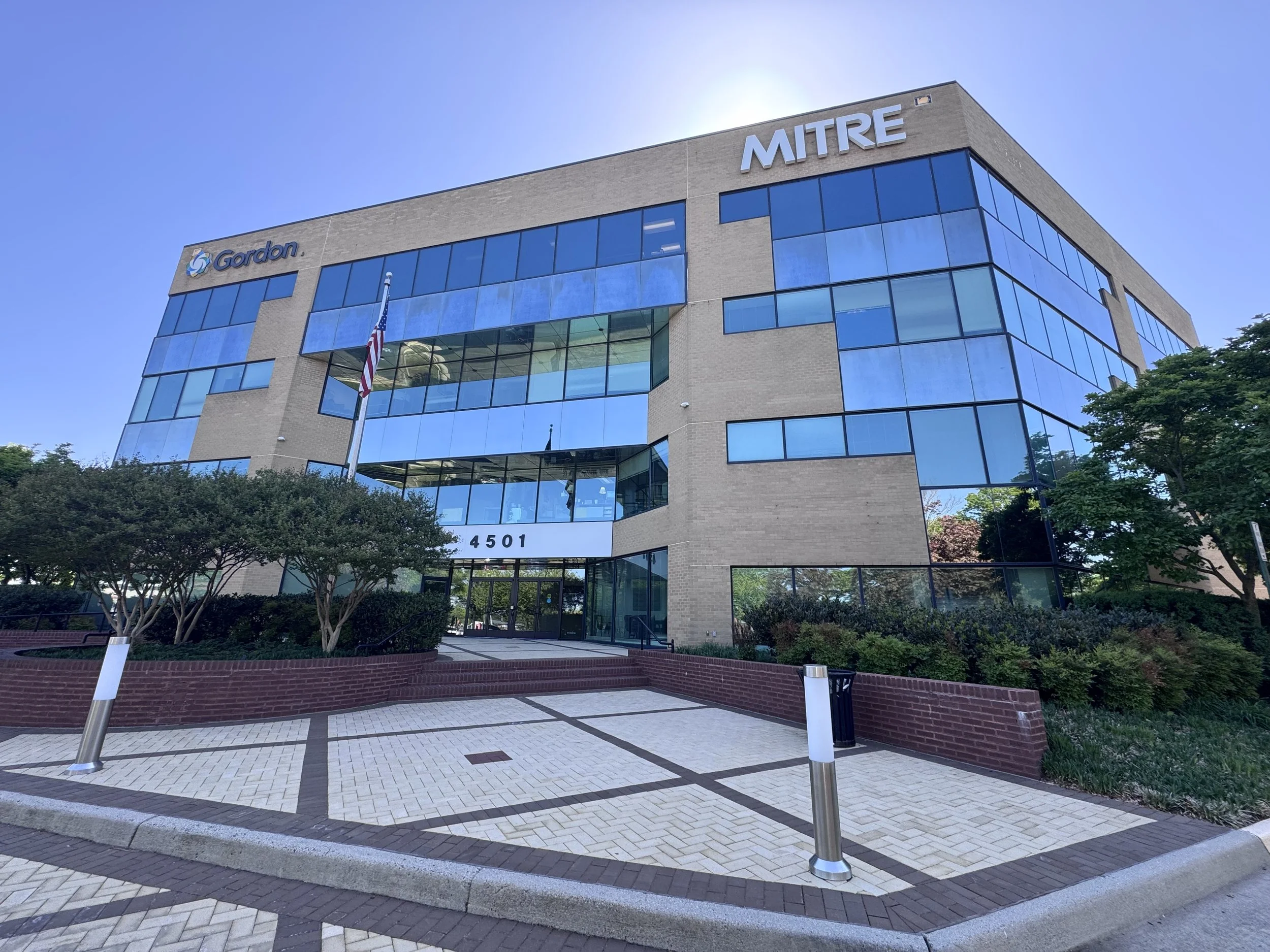 Photo of a modern office building with glass windows, the street number 4501, flags, some trees, and a clear blue sky.
