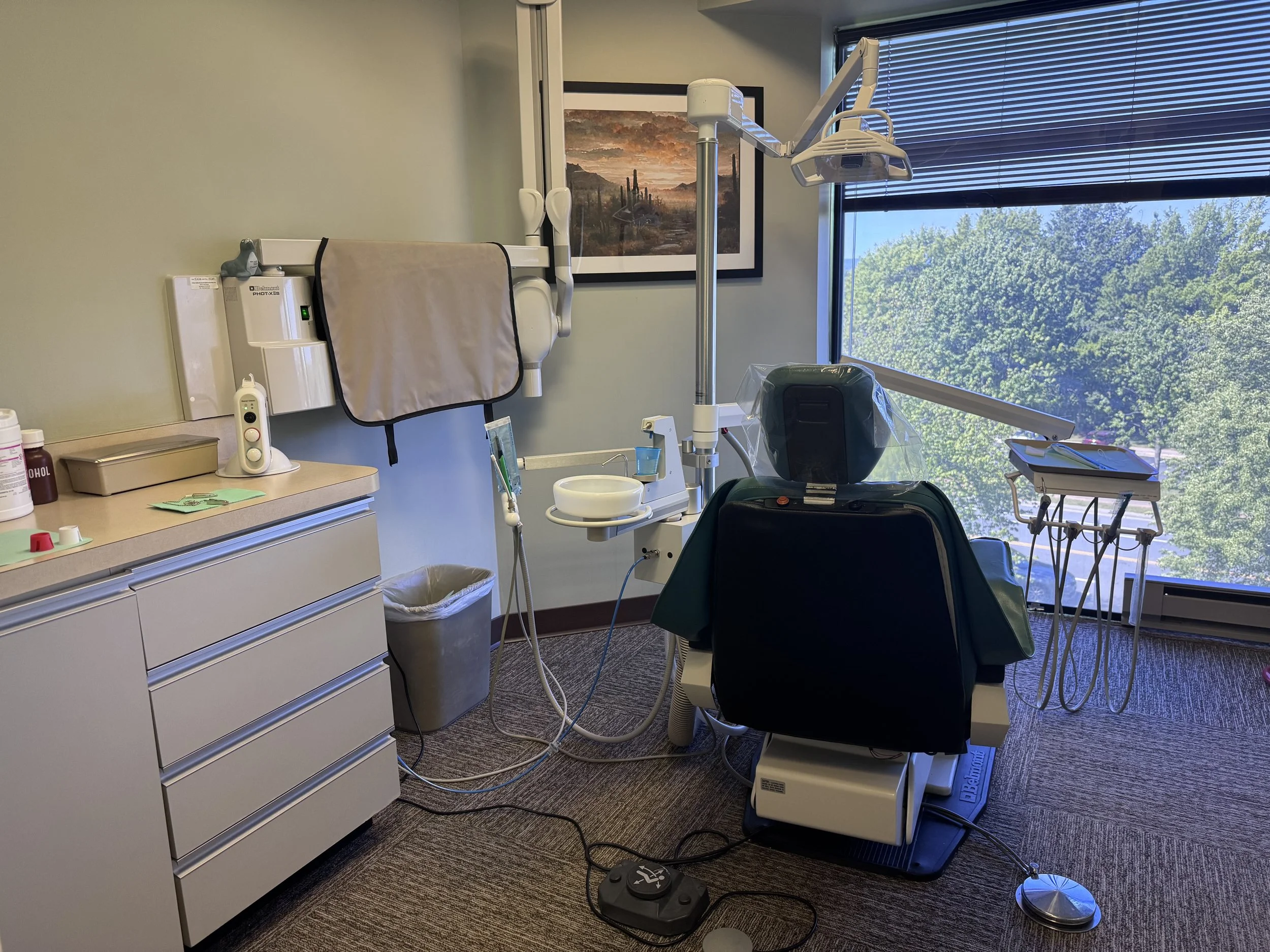 Dental exam room with dental chair, equipment, and a large window showing trees outside.