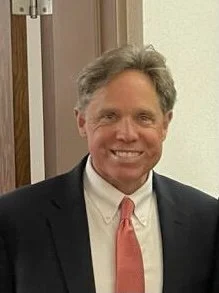 A smiling man with gray hair in a suit and red tie, standing indoors