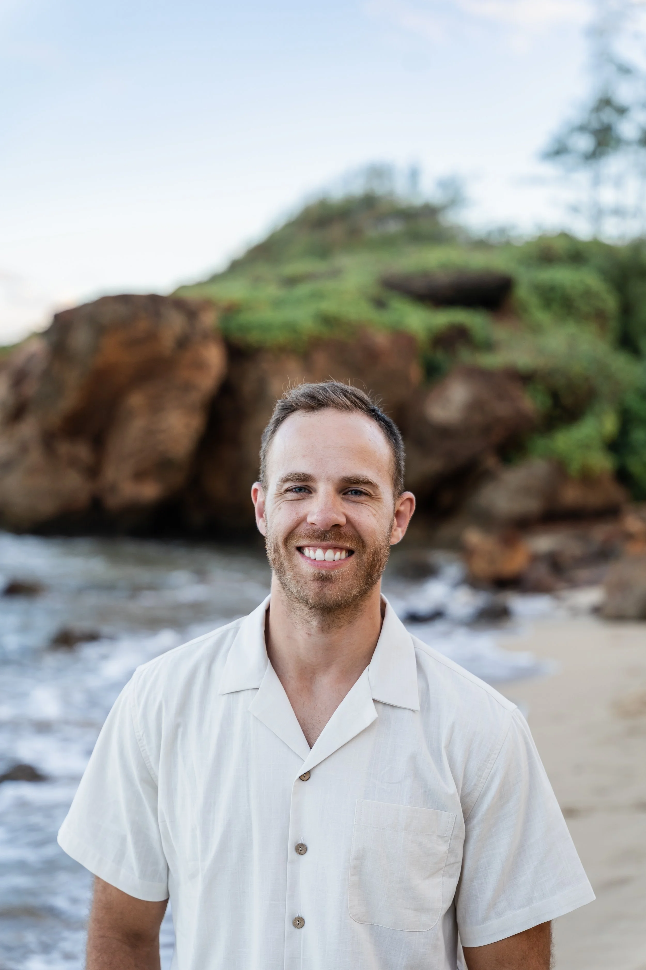 Dr. Jeff smiling at the camera, standing outdoors near the water with rocks and greenery in the background. He has short brown hair and a beard.