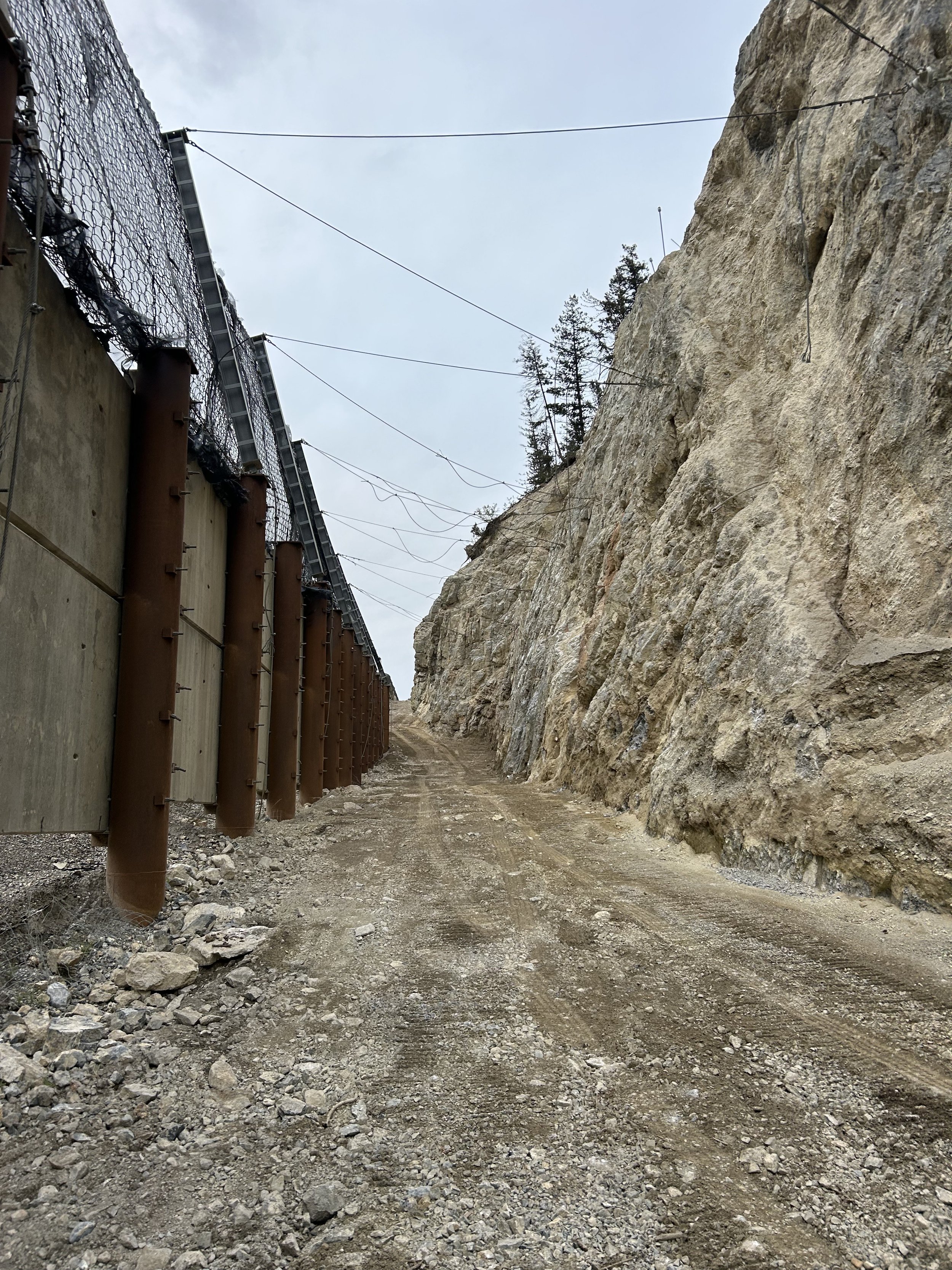 A dirt road with tire tracks running between a steep rocky cliff on the right and a tall metal fence with barbed wire on the left, with trees and an overcast sky overhead.