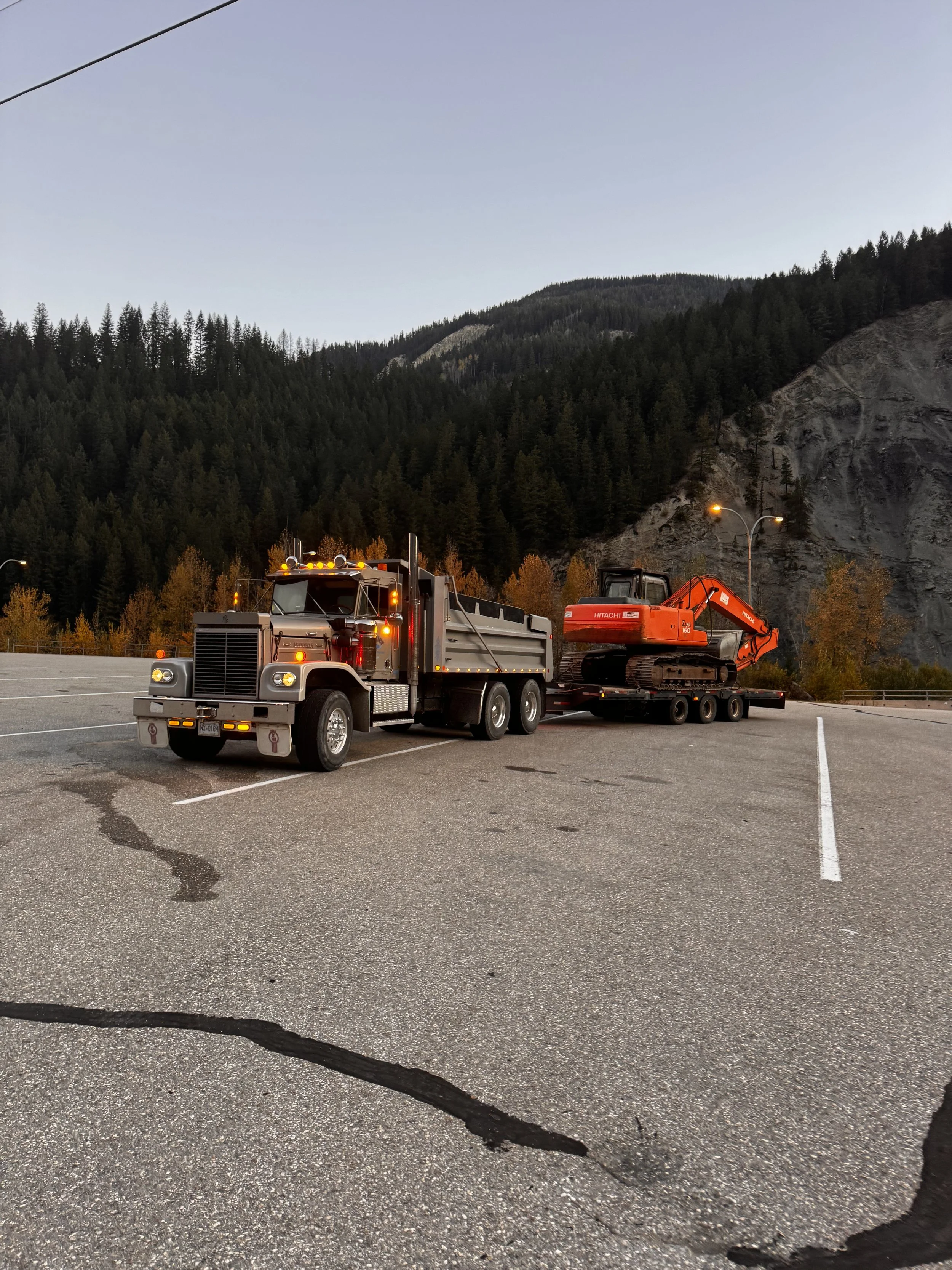 A flatbed truck carrying an orange excavator parked on an empty parking lot with mountains and trees in the background.