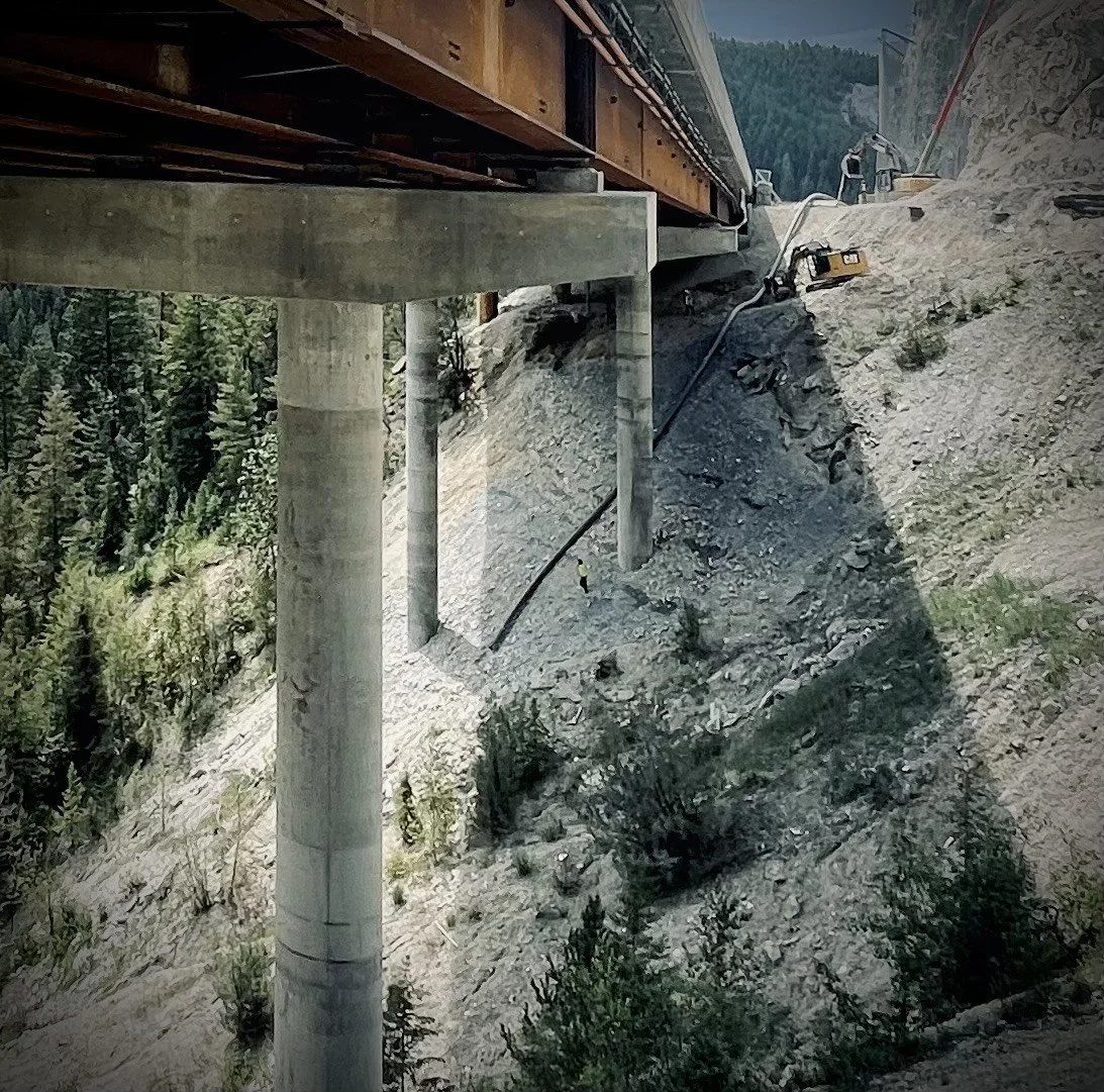 Construction work on a bridge, showing large support pillars and equipment on the rocky hillside.