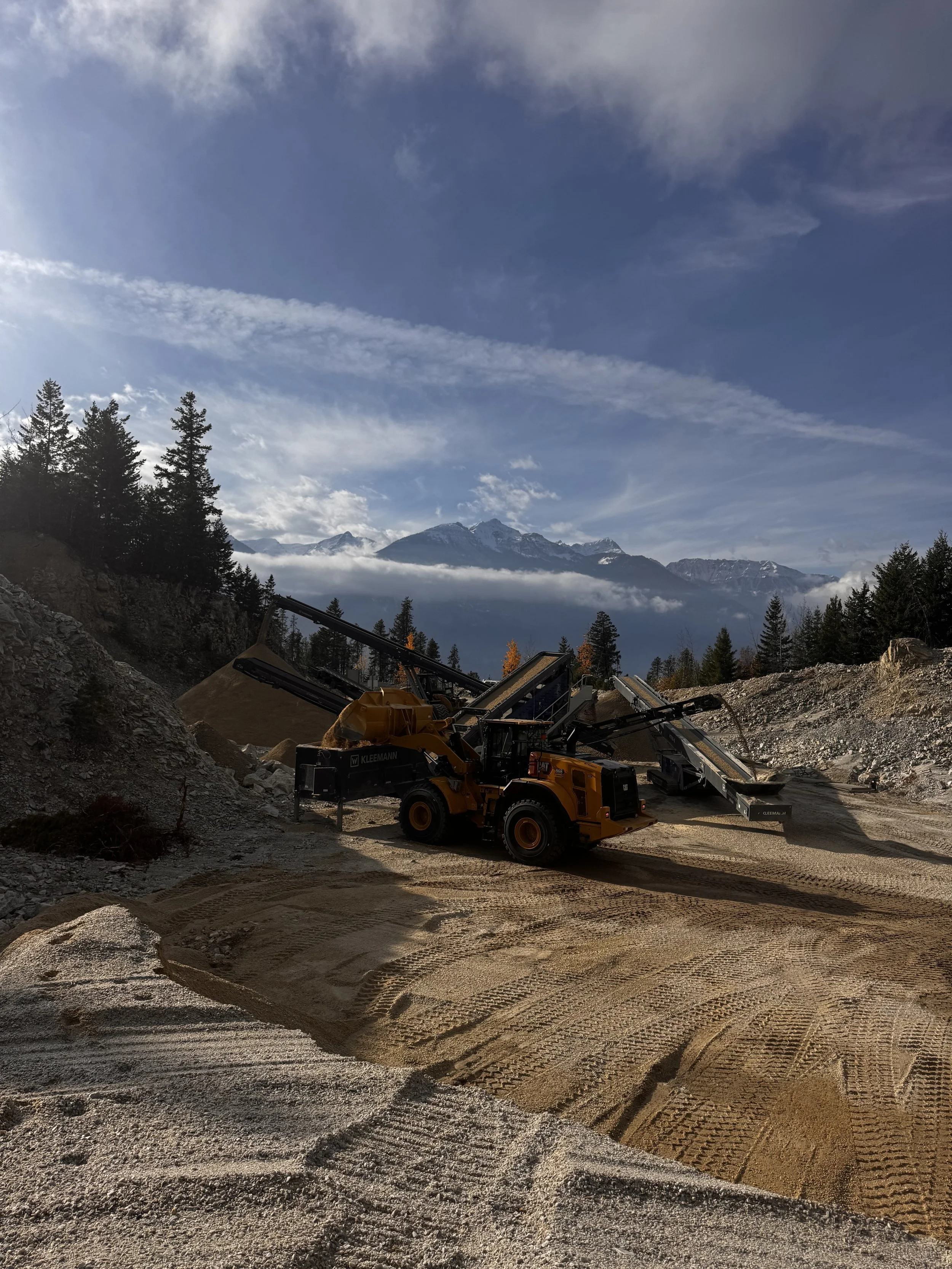 Construction site with a large yellow and black bulldozer moving gravel, surrounded by rocky terrain and forested mountains in the background under a cloudy sky.