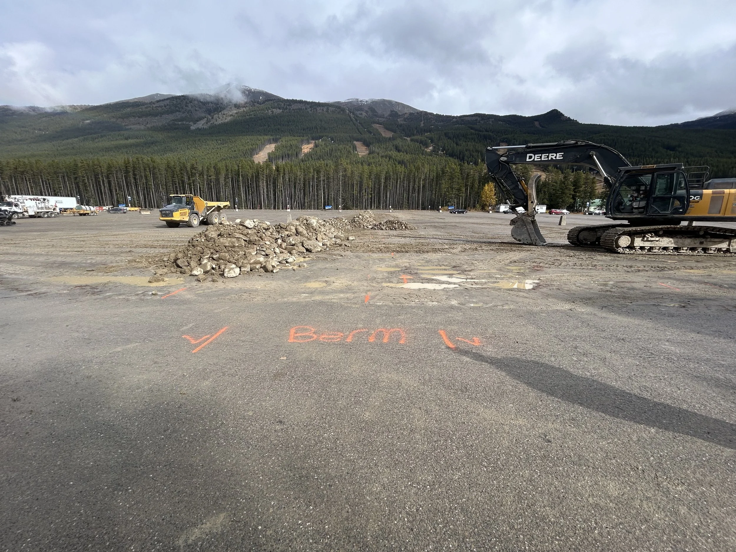Construction site with dirt piles and equipment, including a large excavator marked 'DEERE,' on a dirt lot with mountains and trees in the background, and orange markings on the ground.