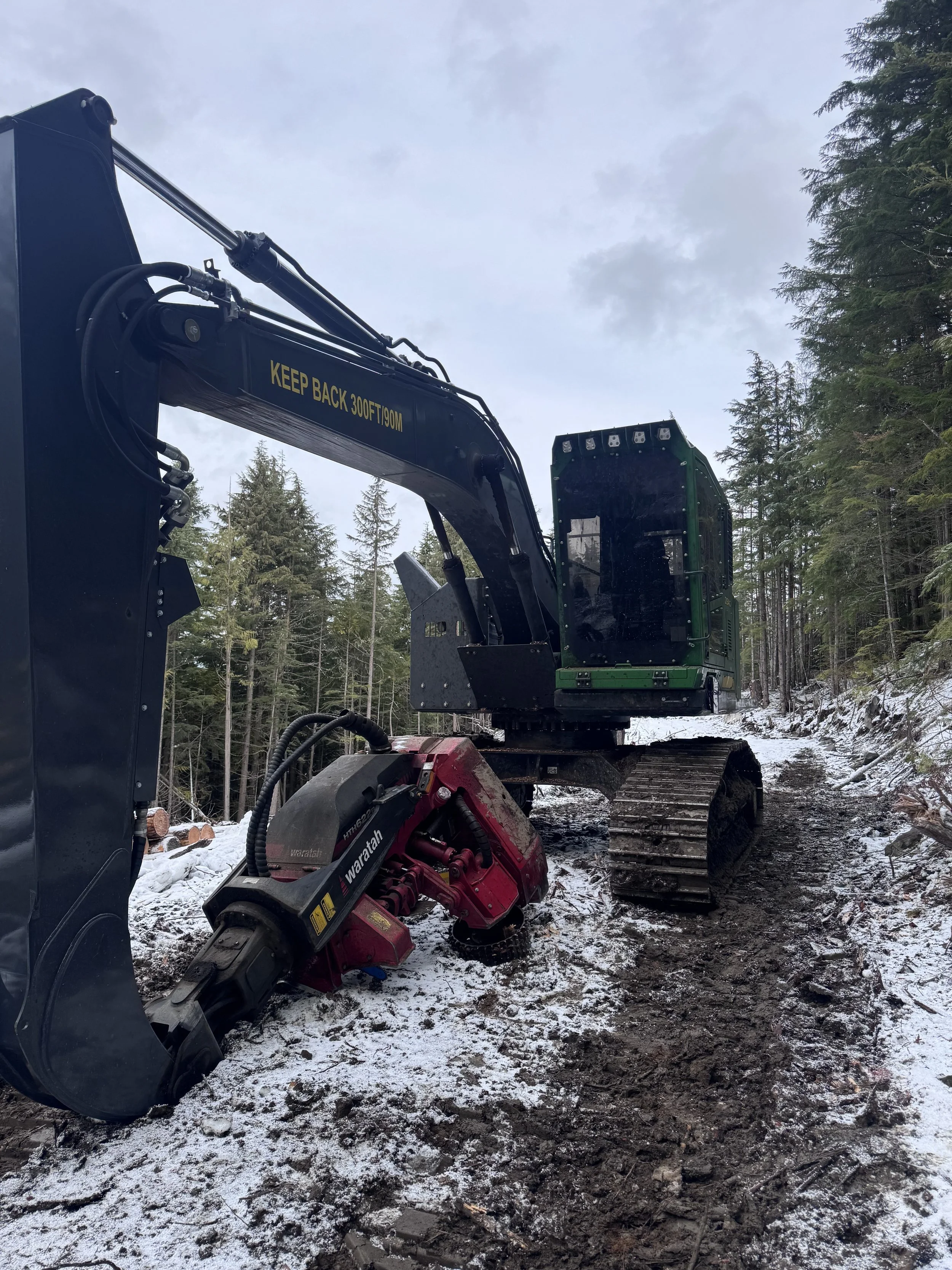 Heavy construction excavator on snow-covered dirt path in a forest, with tall trees and cloudy sky in the background.