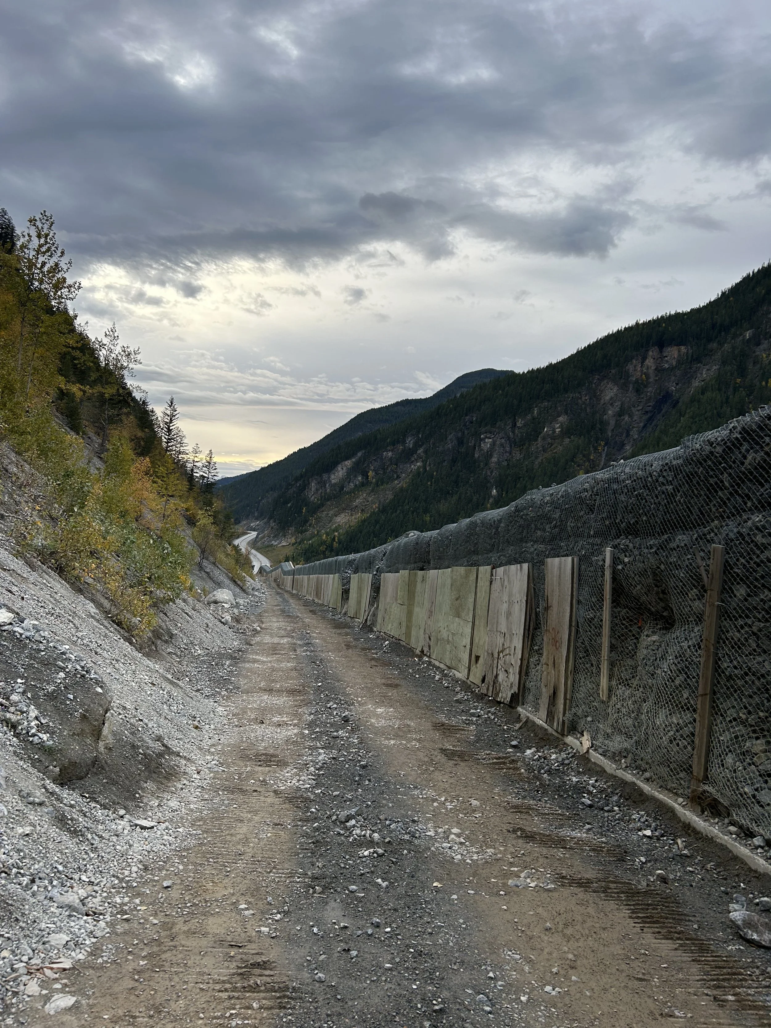 A dirt mountain road with a protective fence on the right side and yellow and green foliage on the left, under a cloudy sky.