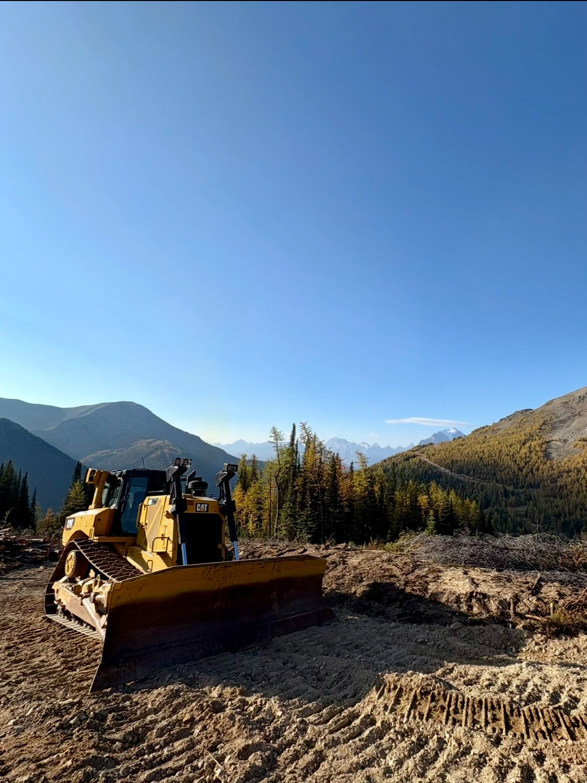 A yellow bulldozer on dirt with mountains and trees in the background under a clear blue sky.