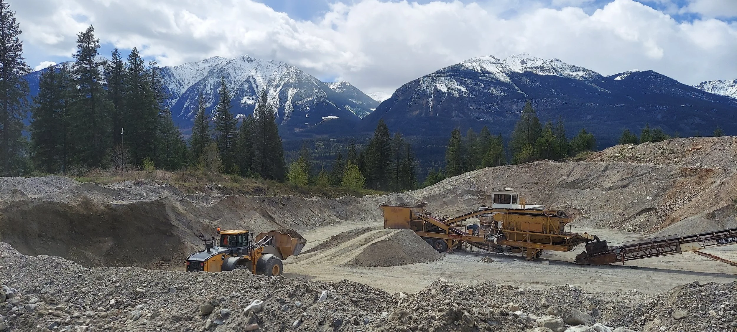 Construction site with heavy machinery operating on rocky terrain, mountains with snow-covered peaks and a forest of pine trees in the background.