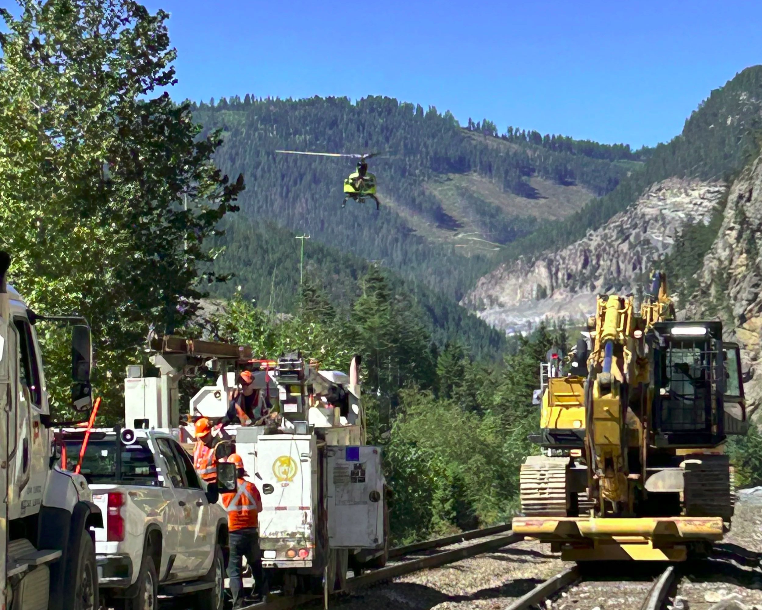 A workers at a train maintenance site in a forested mountainous area, with a helicopter flying overhead and construction equipment on the railway tracks.