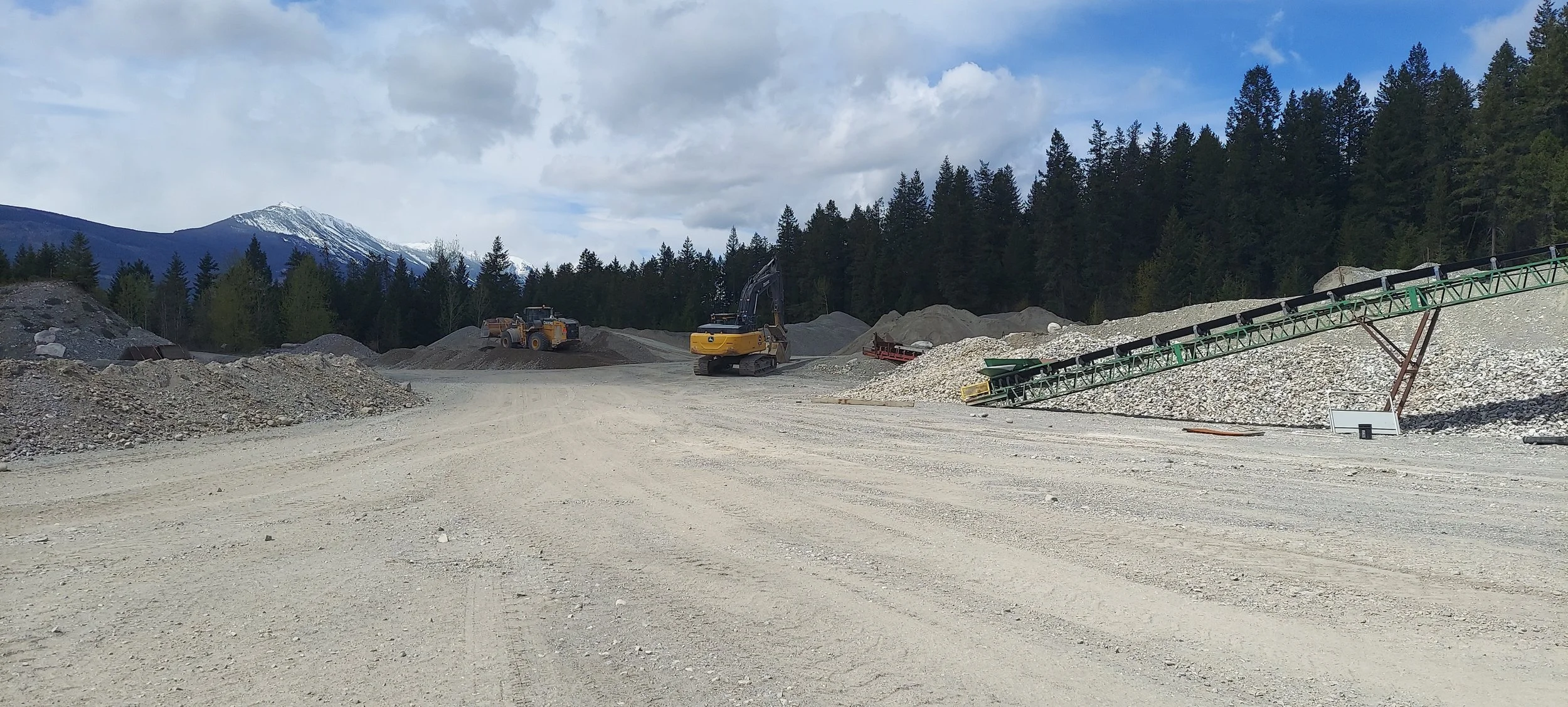 Construction site with gravel piles, a conveyor belt, and heavy machinery in front of a forest and snowy mountain in the background.