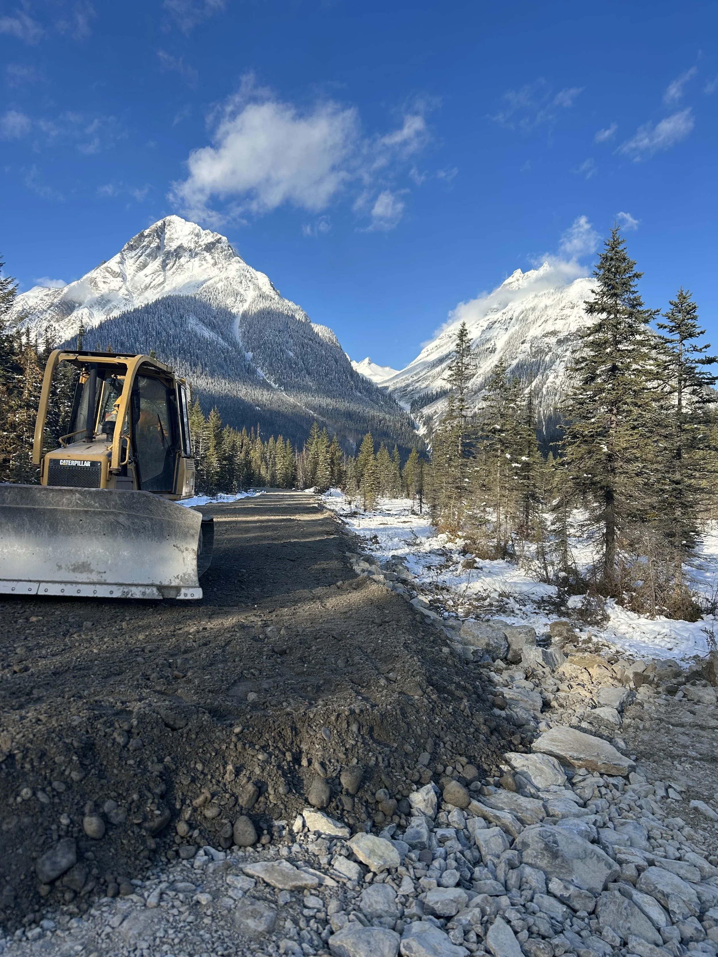 A dirt road being constructed with a bulldozer on a snowy mountain landscape with pine trees and snow-capped peaks in the background.