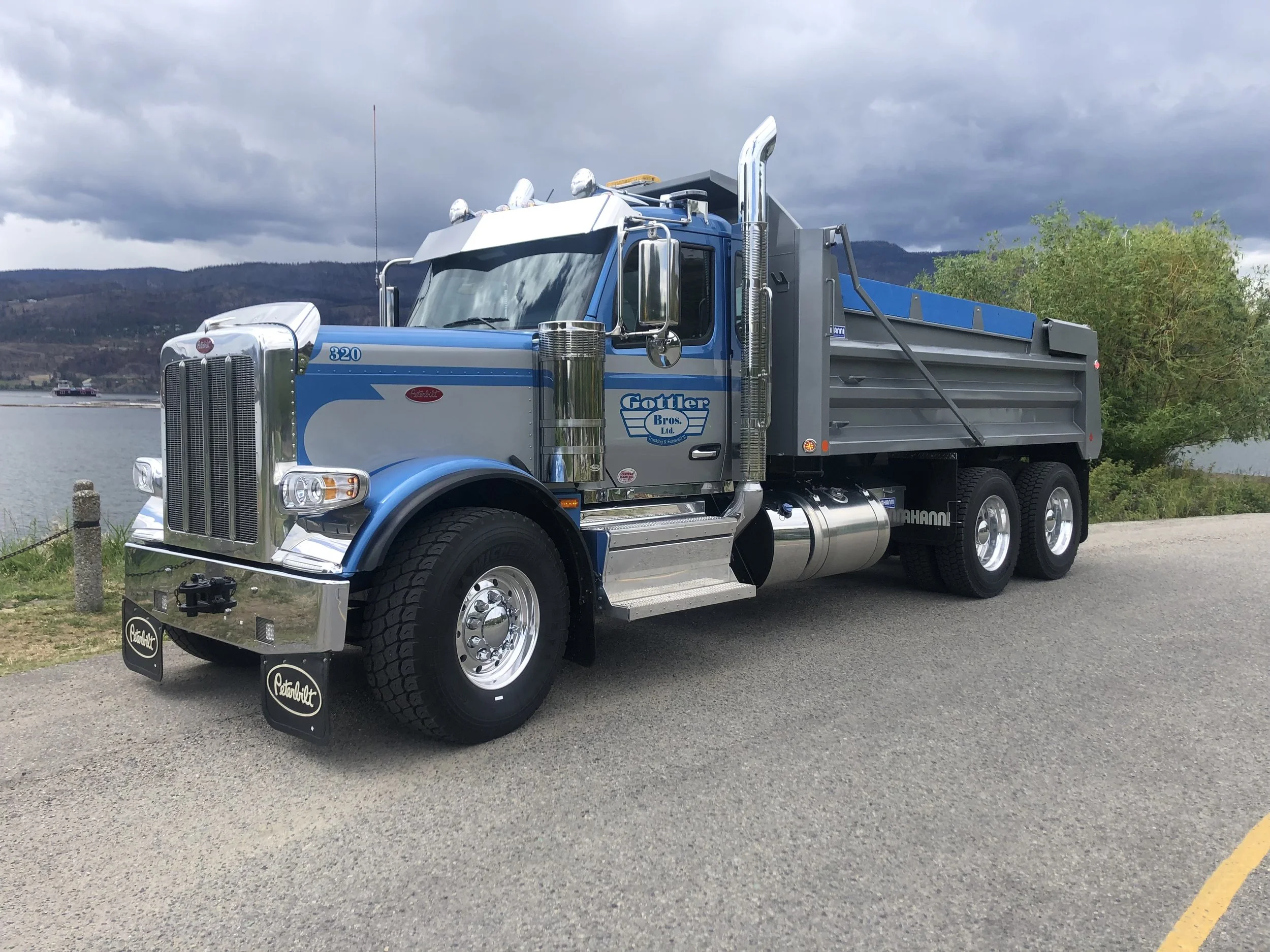 A vintage Peterbilt dump truck parked on a scenic roadside near water, with mountains and cloudy sky in the background.