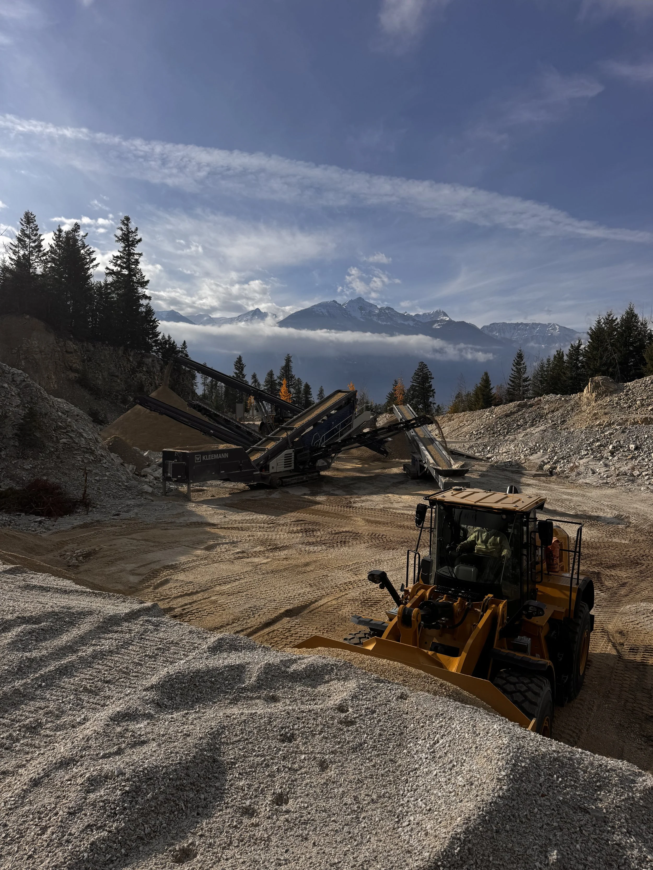 Construction site with a bulldozer and a conveyor belt system on uneven ground, surrounded by trees and mountains in the background under a partly cloudy sky.