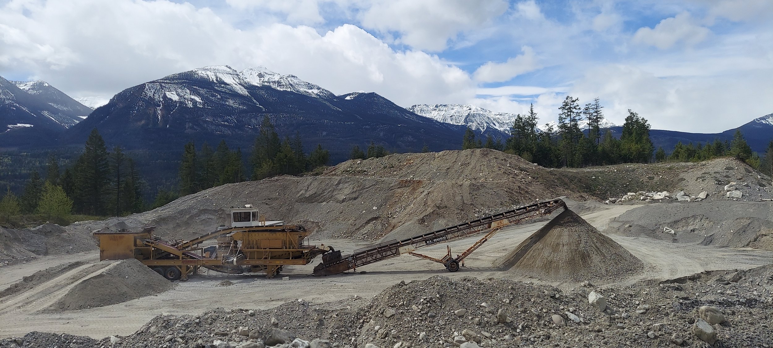 A gravel quarry with machinery processing rocks, set against a backdrop of forested mountains with snow-capped peaks and a partly cloudy sky.