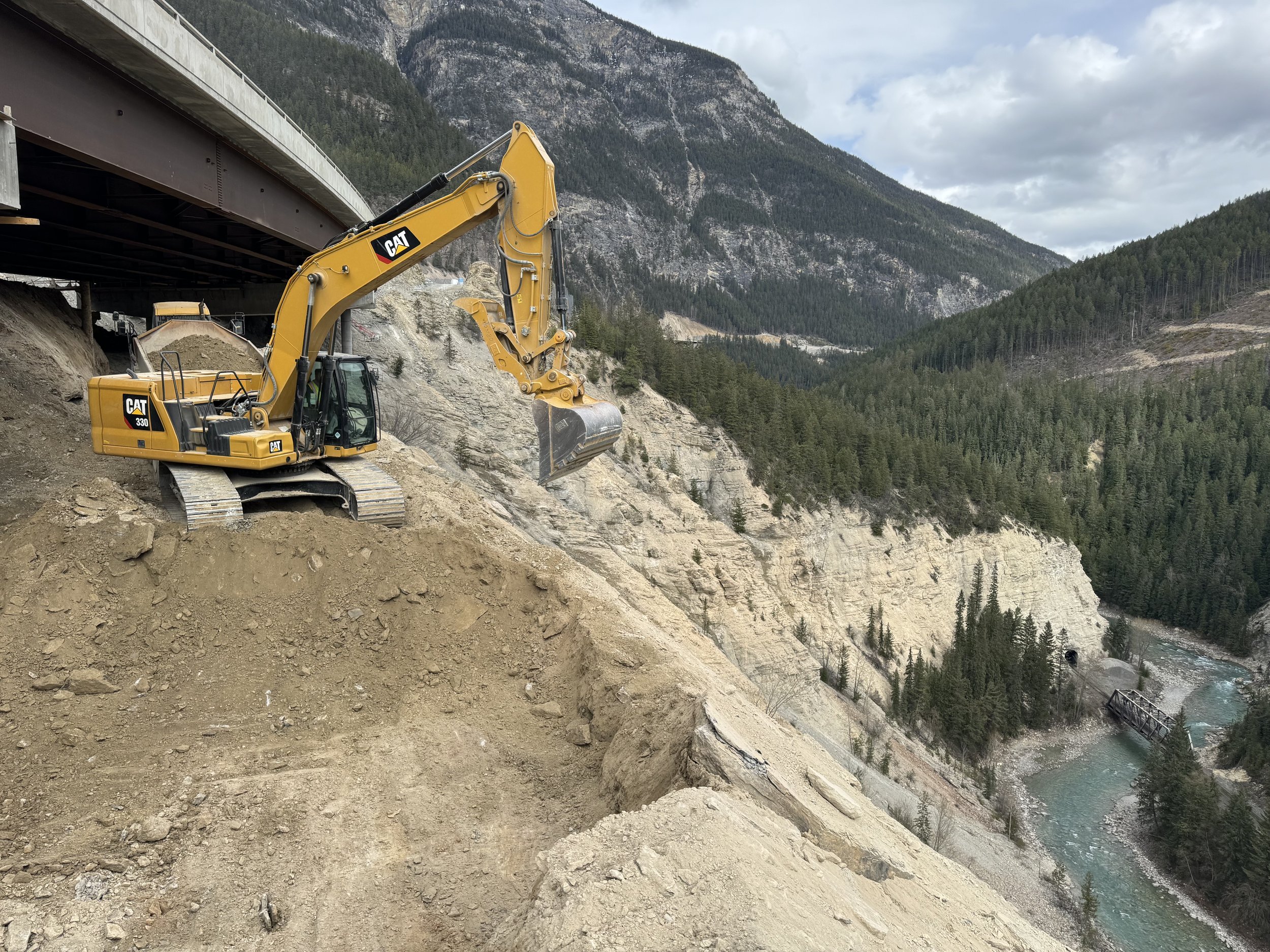 A yellow excavator working on a construction site on the side of a mountain, overlooking a river and forested valleys, with a bridge in the distance and a cloudy sky overhead.