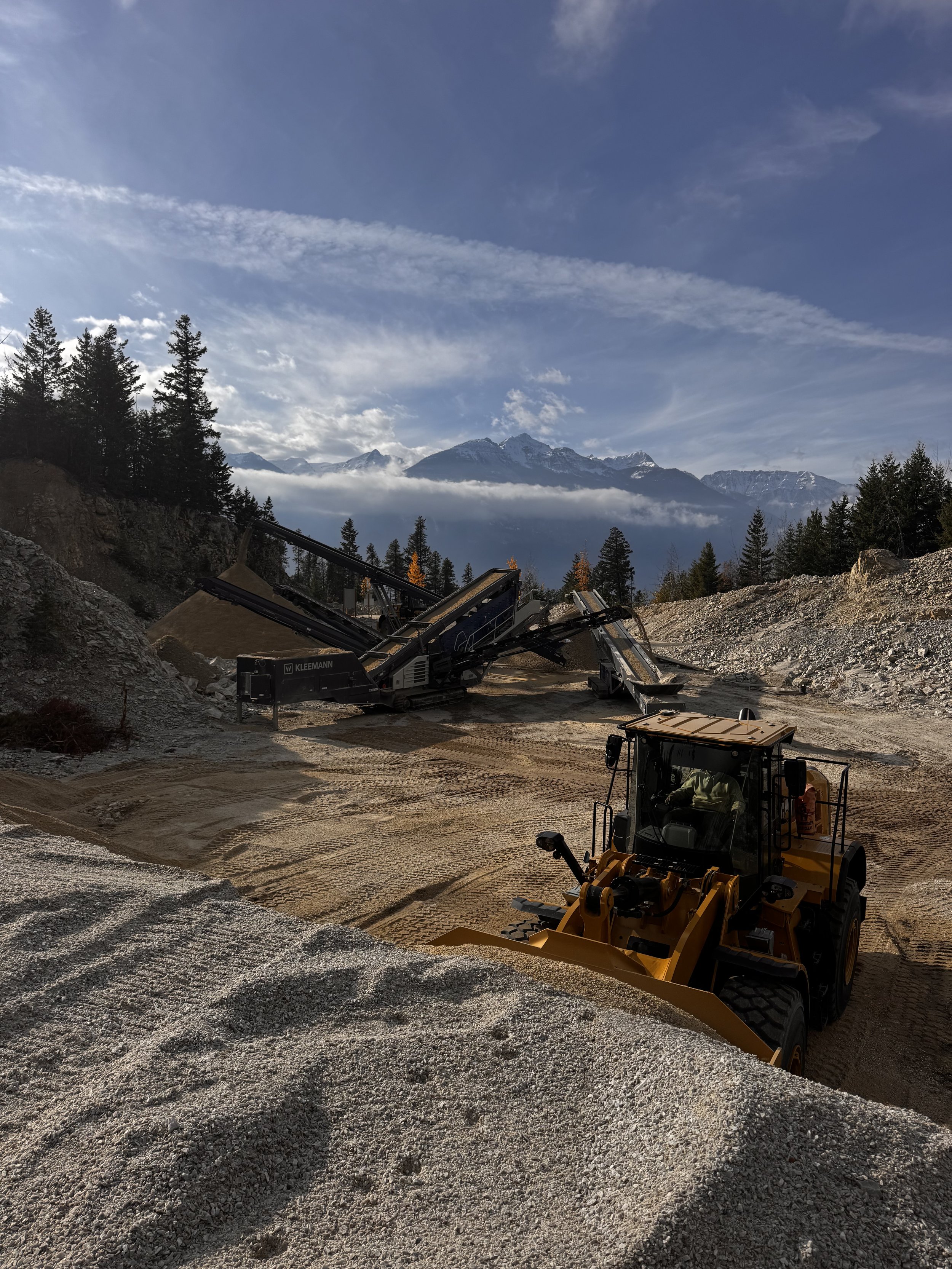 Construction site with a yellow front loader and a gravel screener machine, set against mountain peaks and a partly cloudy sky.
