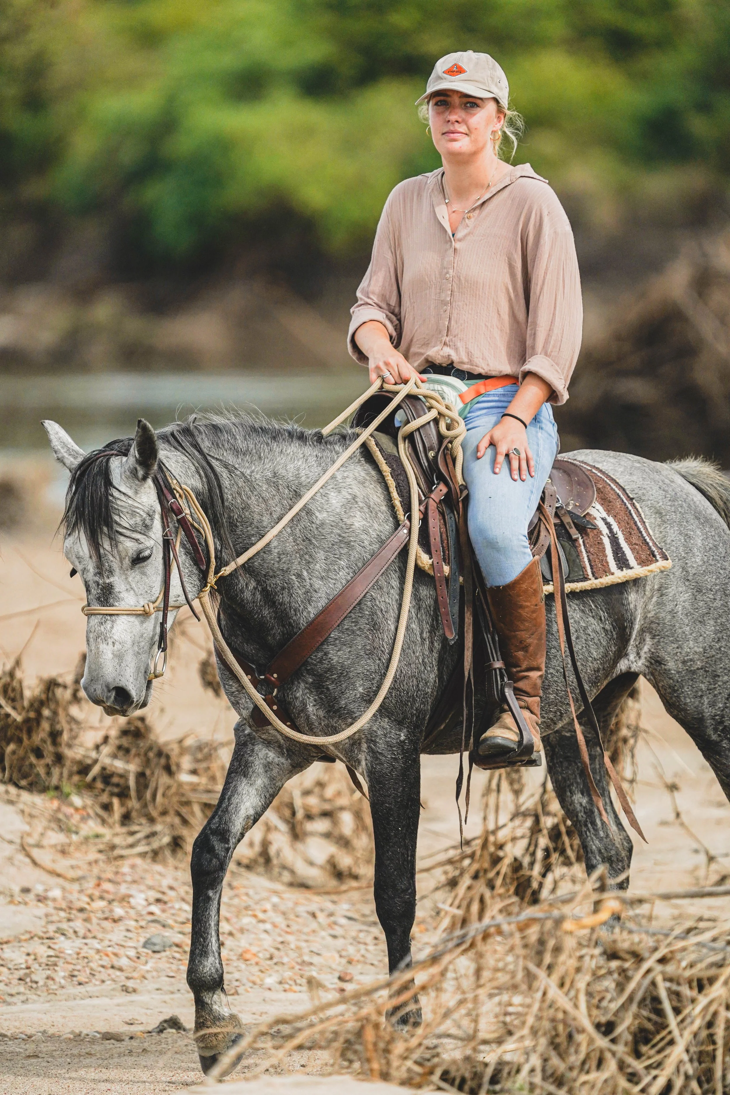 A woman riding a gray horse along a natural outdoor trail with greenery in the background. She is wearing a beige cap, a light brown long-sleeve shirt, blue jeans, and brown boots.