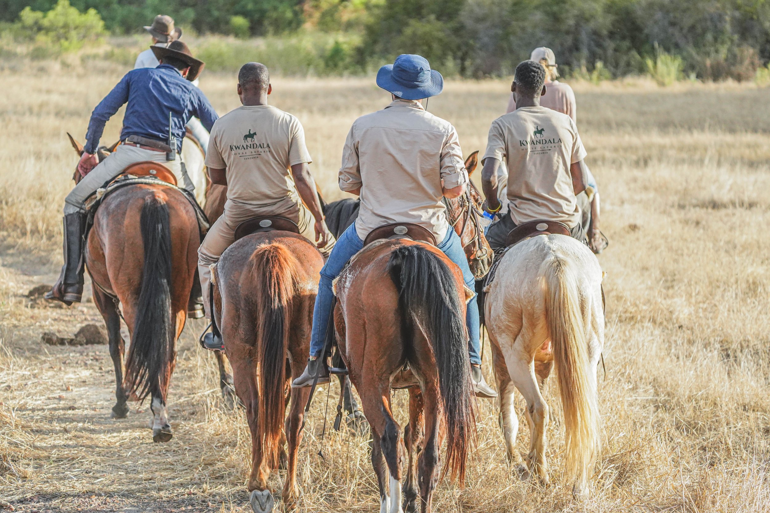 Group of people riding horses on a dirt trail through a grassy field with trees in the background. Some individuals are wearing hats, and two of them are wearing shirts with 