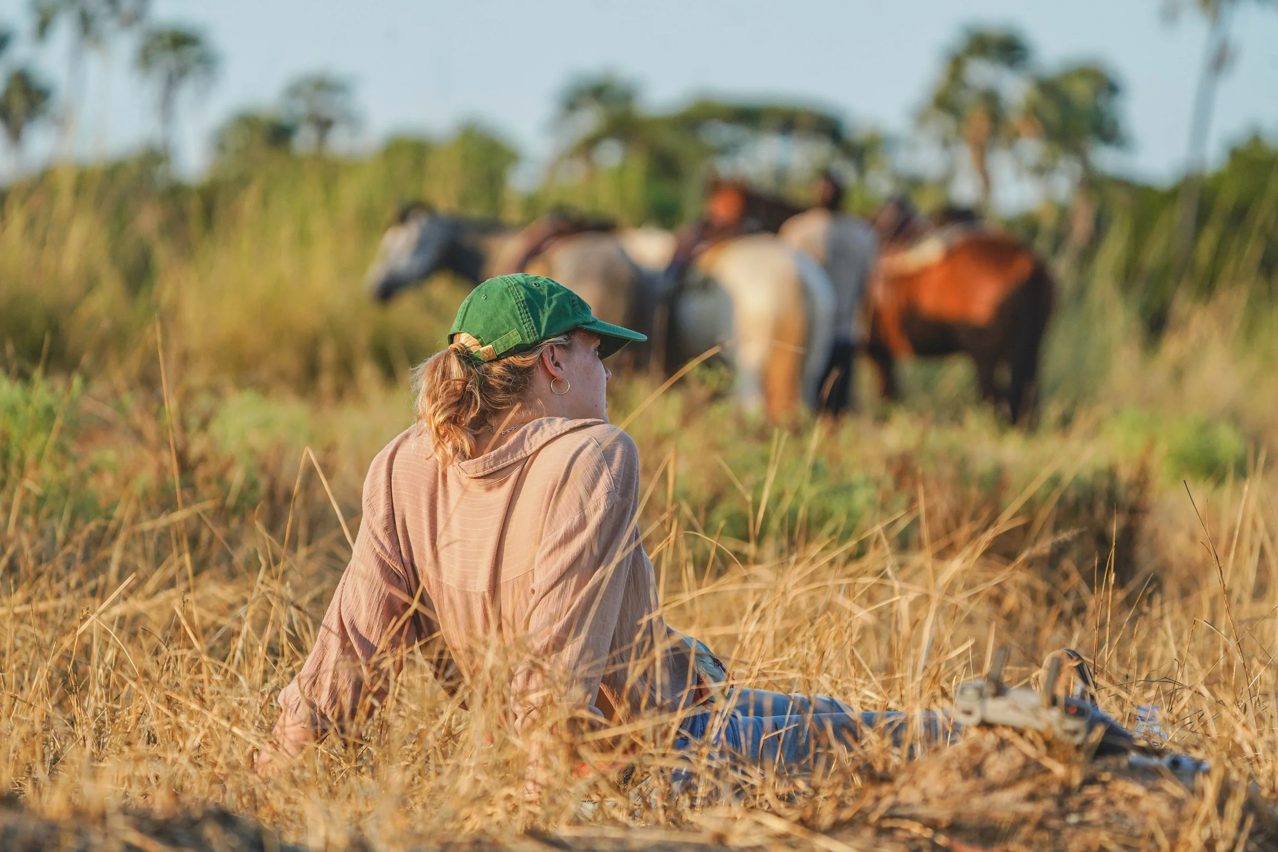 A woman with a green cap sitting on dry grass near horses in a field, with horses in the background. a moments rest on a horse safari in tanzania