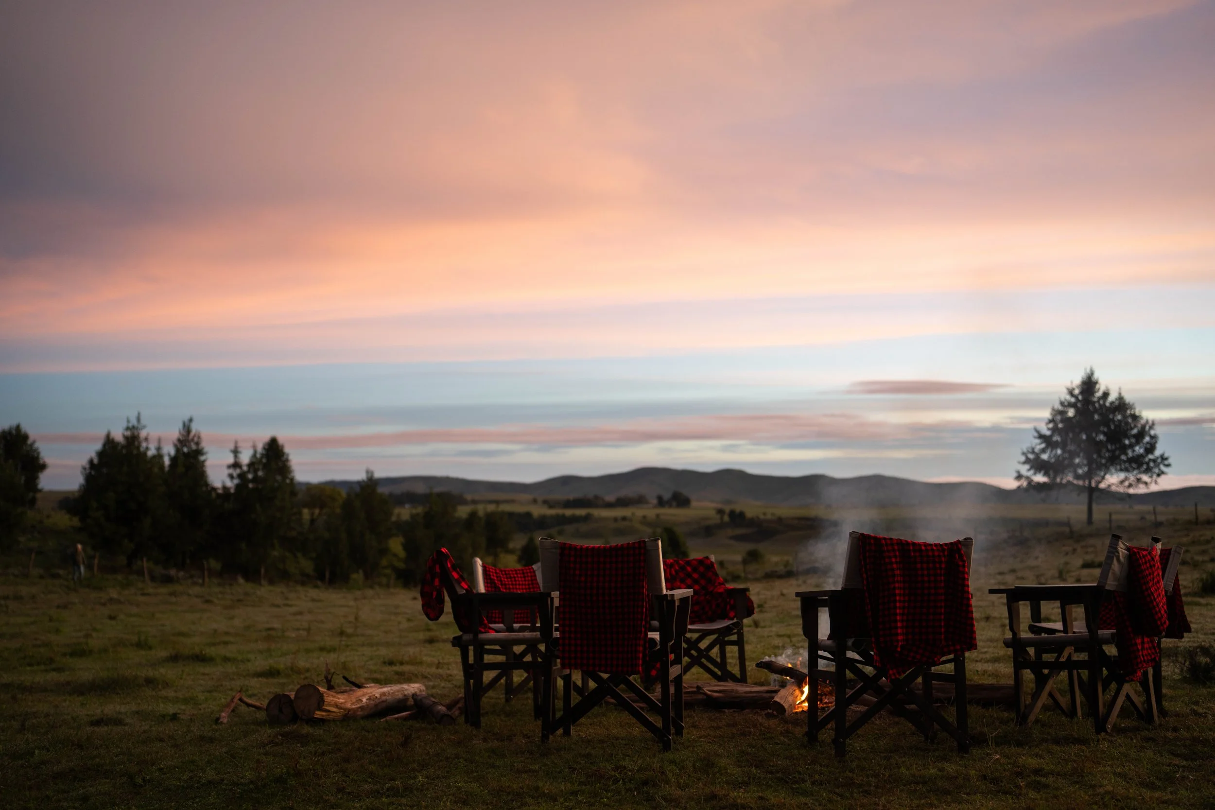 Outdoor scene at sunset with several chairs arranged around a small fire, rolling hills in the background, trees, and a sky with pink and purple clouds. Mobile camp for our riding safaris