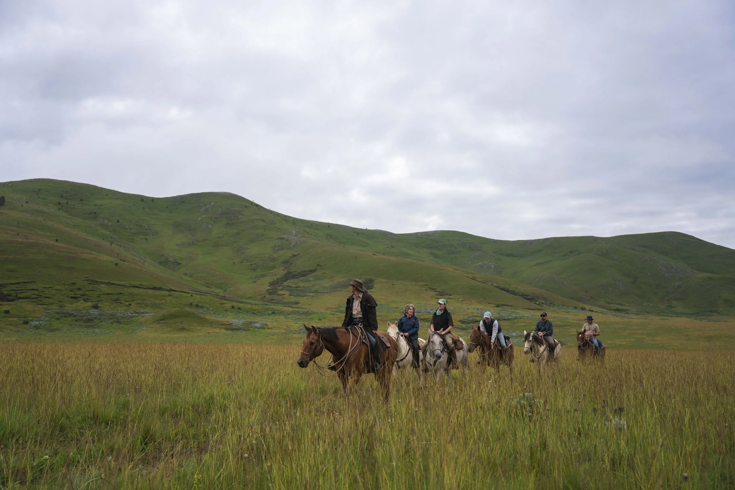 A group of people horseback riding through a green grassy field with rolling hills and cloudy sky in the background.
