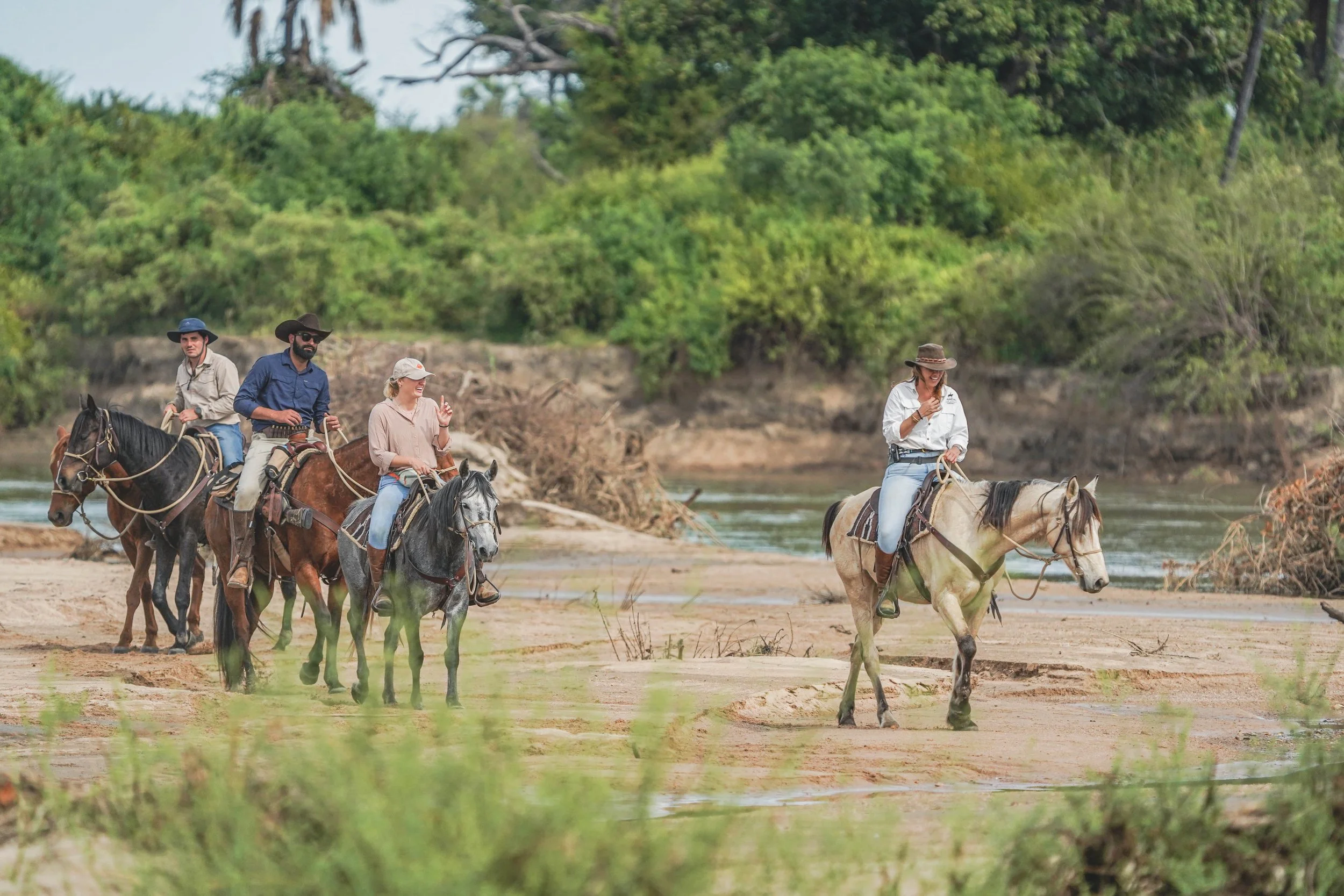 Four people horseback riding along a riverbank surrounded by greenery and trees.