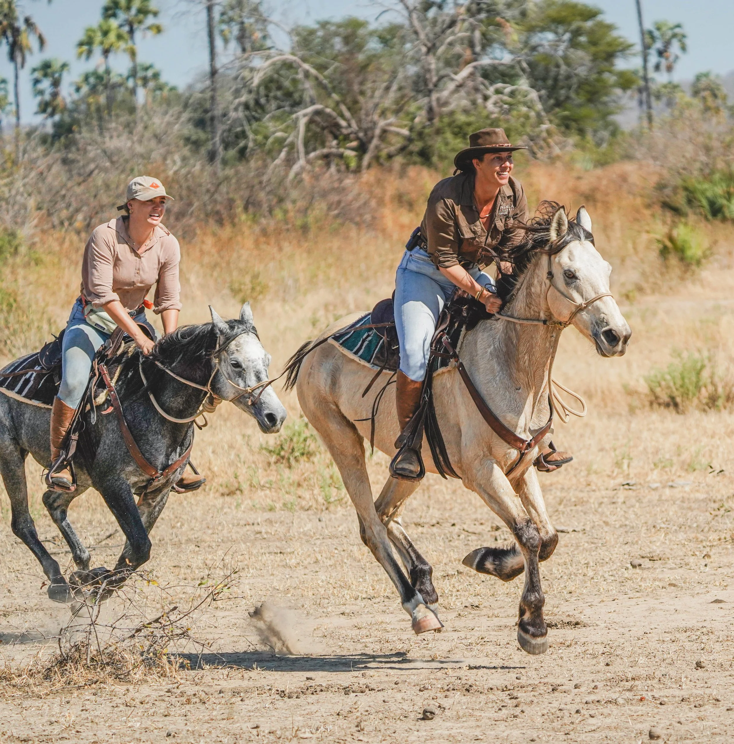 Two women riding horses through a dry, grassy landscape with trees in the background.