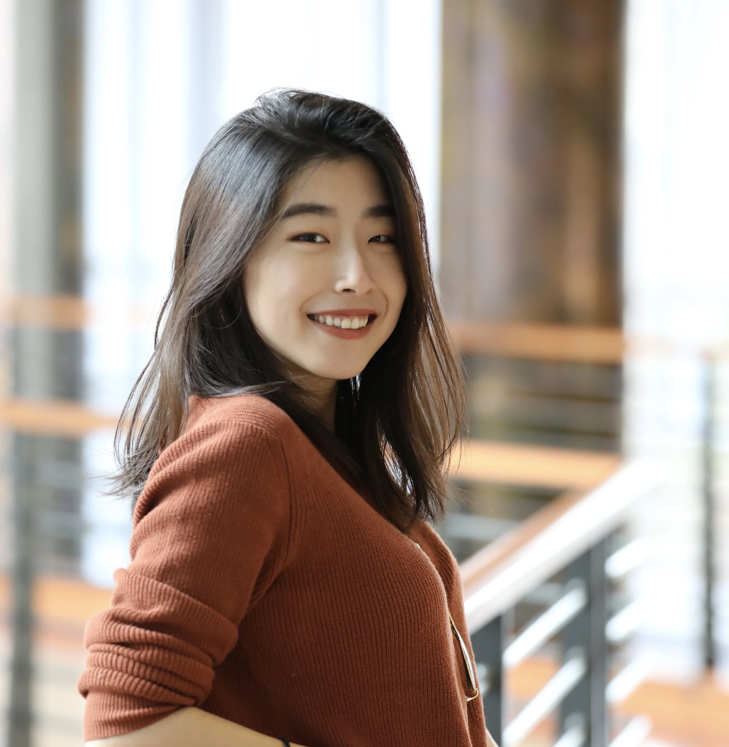 A smiling Asian woman with shoulder-length dark hair, wearing a rust-colored sweater, standing indoors near a staircase with blurred background.