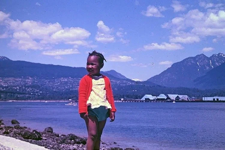 Portrait of Myra Lee Virgil, memoir writer and essayist based in Bermuda, as a young girl. She has braided hair and wears a red and yellow jacket and shorts. She is smiling as she overlooks a large body of water with mountains in Canada.