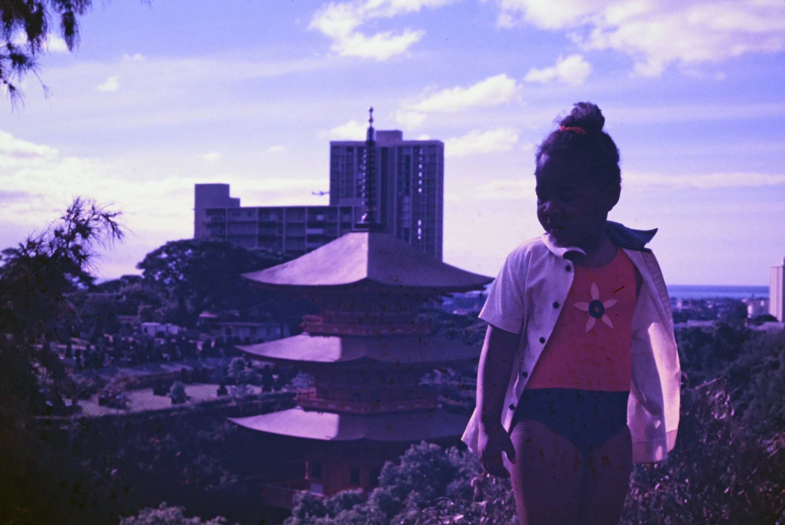 A young girl standing outdoors with a cityscape, including a traditional pagoda-style building and tall modern buildings, in the background on a sunny day with partly cloudy sky.