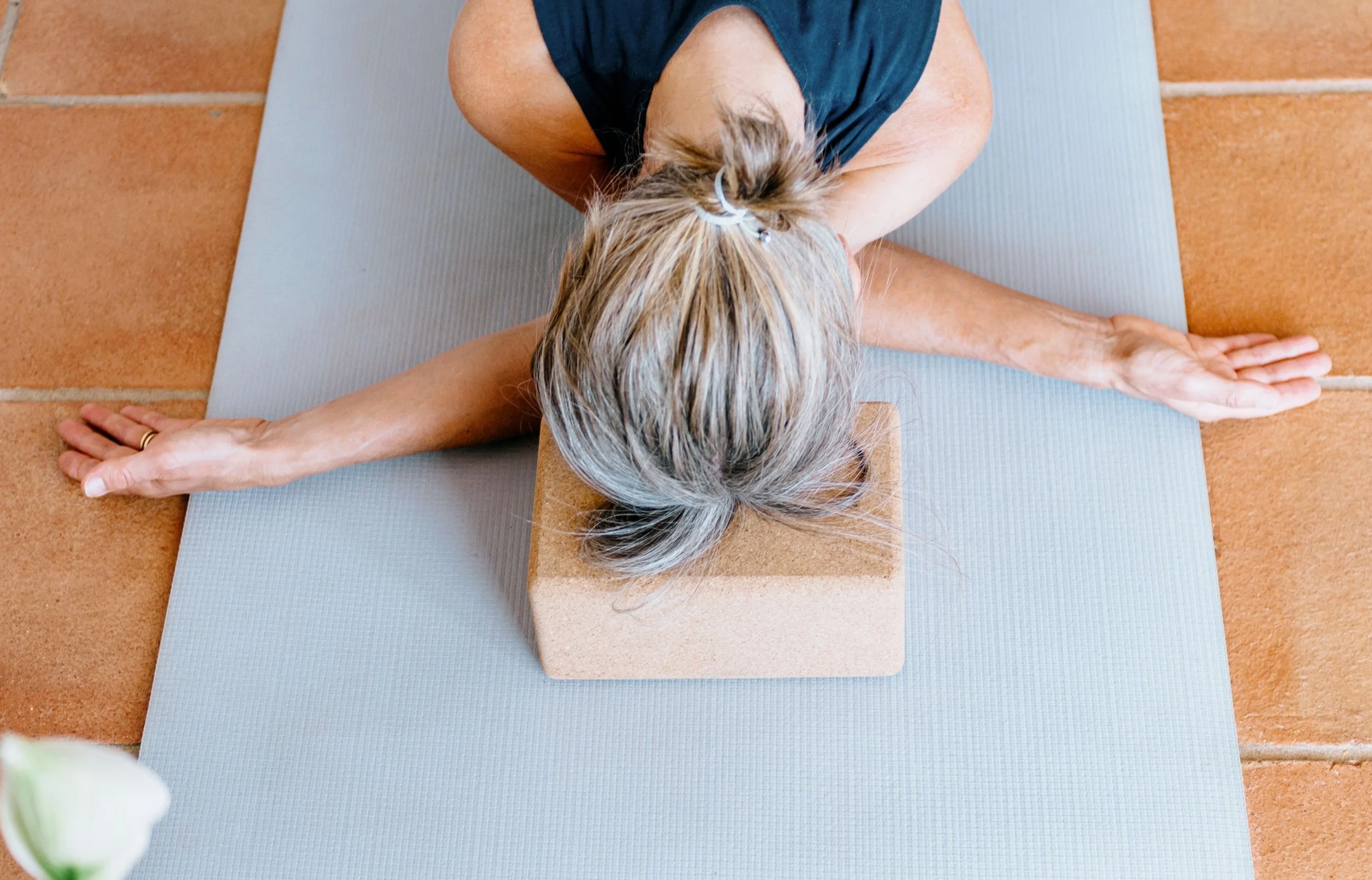 Overhead view of a person practicing yin yoga, laying face down on a mat with arms crossed. There is a cork block under her head on the floor.