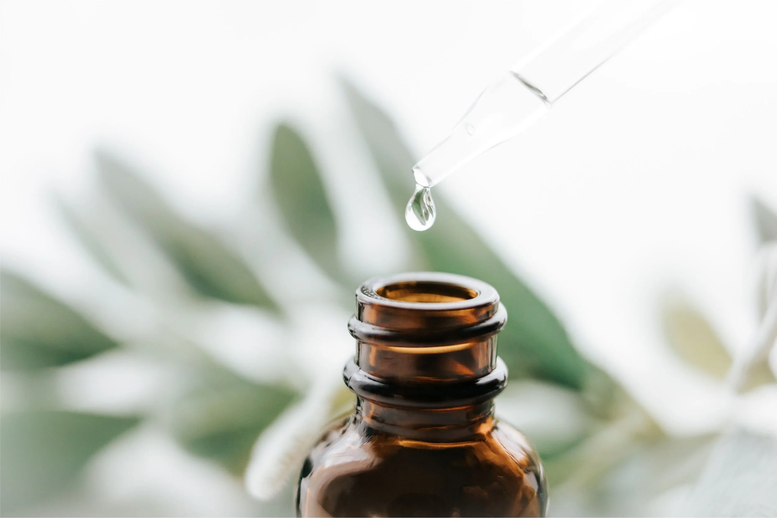 Close-up of a glass essential oil dropper releasing a droplet into an amber glass bottle, with greenery in the blurred background.