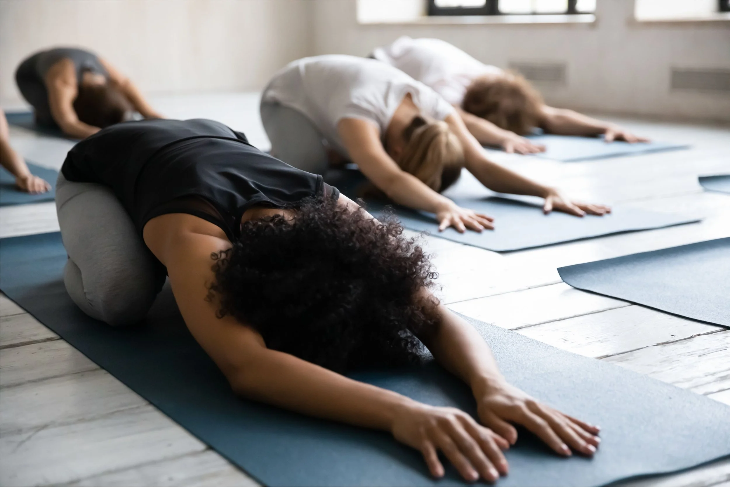 Group of women practicing yoga in child's pose on mats in a well-lit studio.