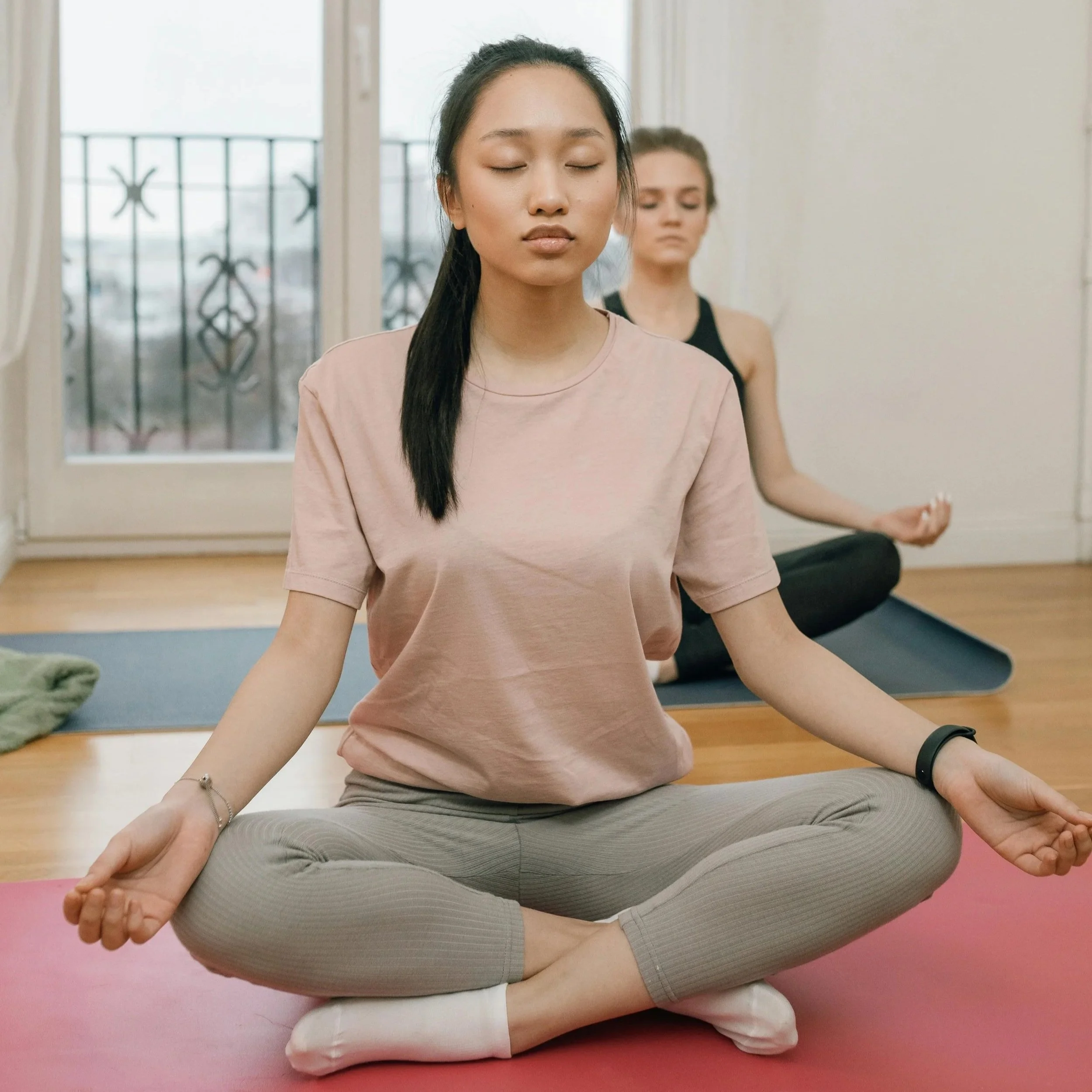 A woman practicing meditation indoors, sitting cross-legged on a yoga mat with closed eyes.