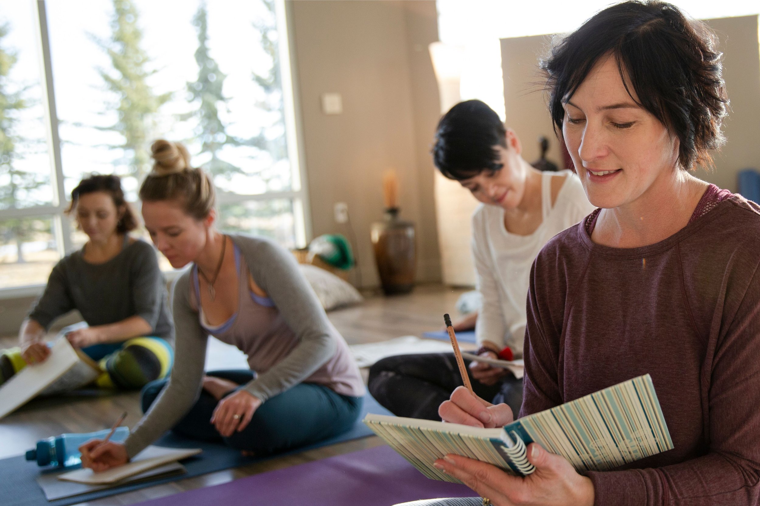 Group of women sitting on the floor in a bright room with large windows, engaging in a reading or writing activity, with notebooks and water bottles in front of them.