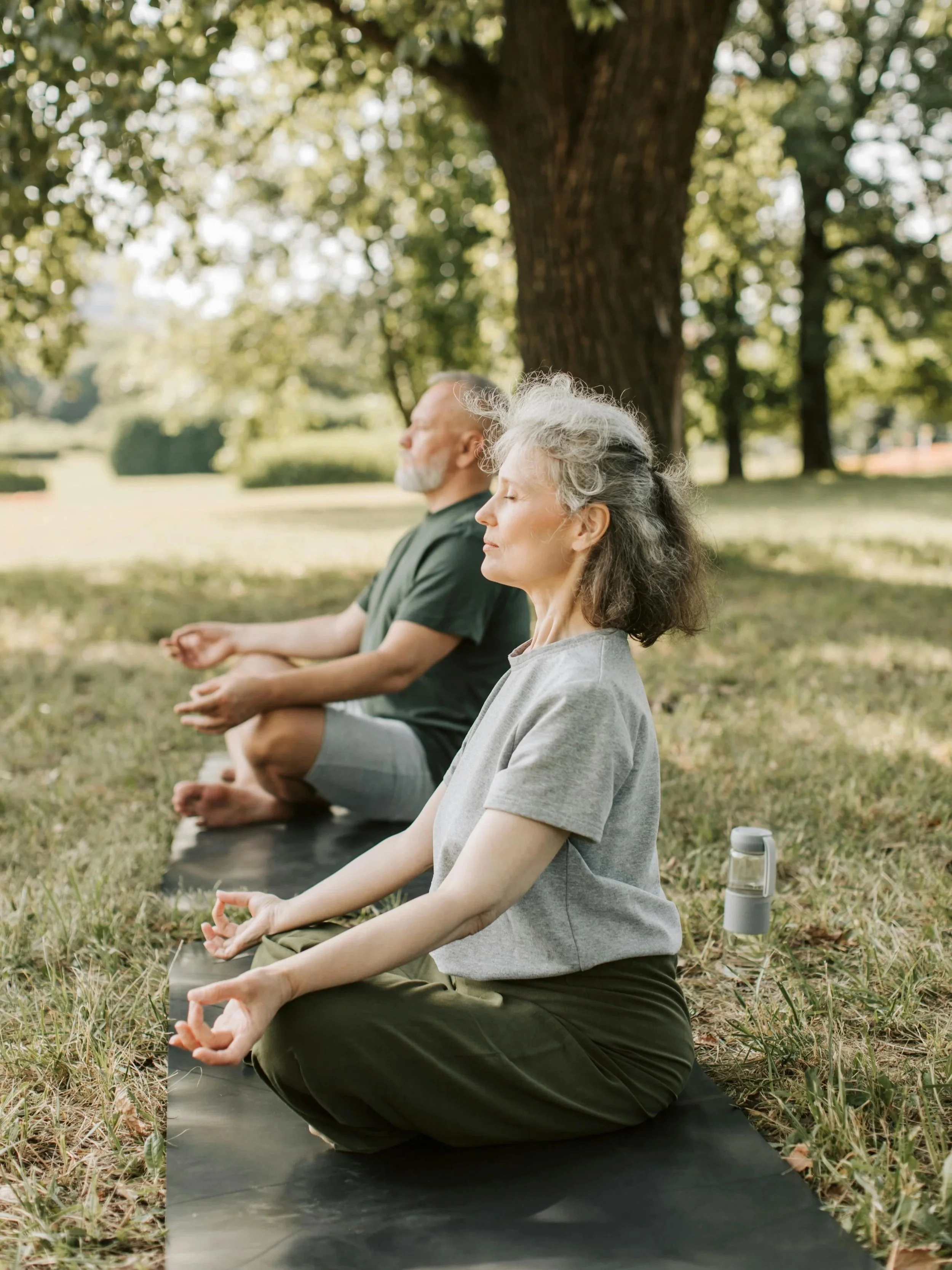 Older man and woman practicing meditation outdoors under a tree, sitting on a black yoga mat with eyes closed and hands in a mudra position.