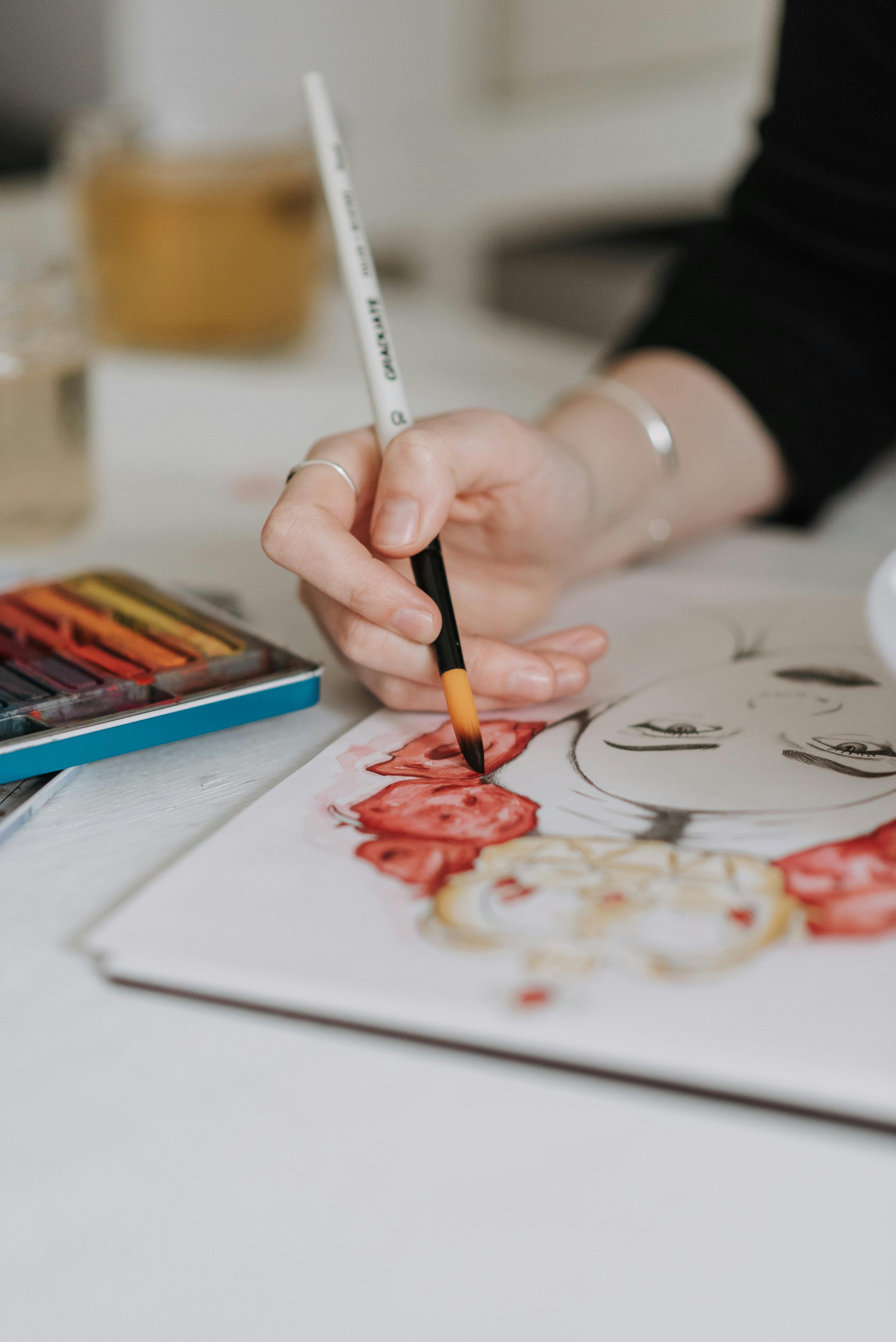 A person is painting on paper with a watercolor brush, creating a floral design with red and yellow colors.
