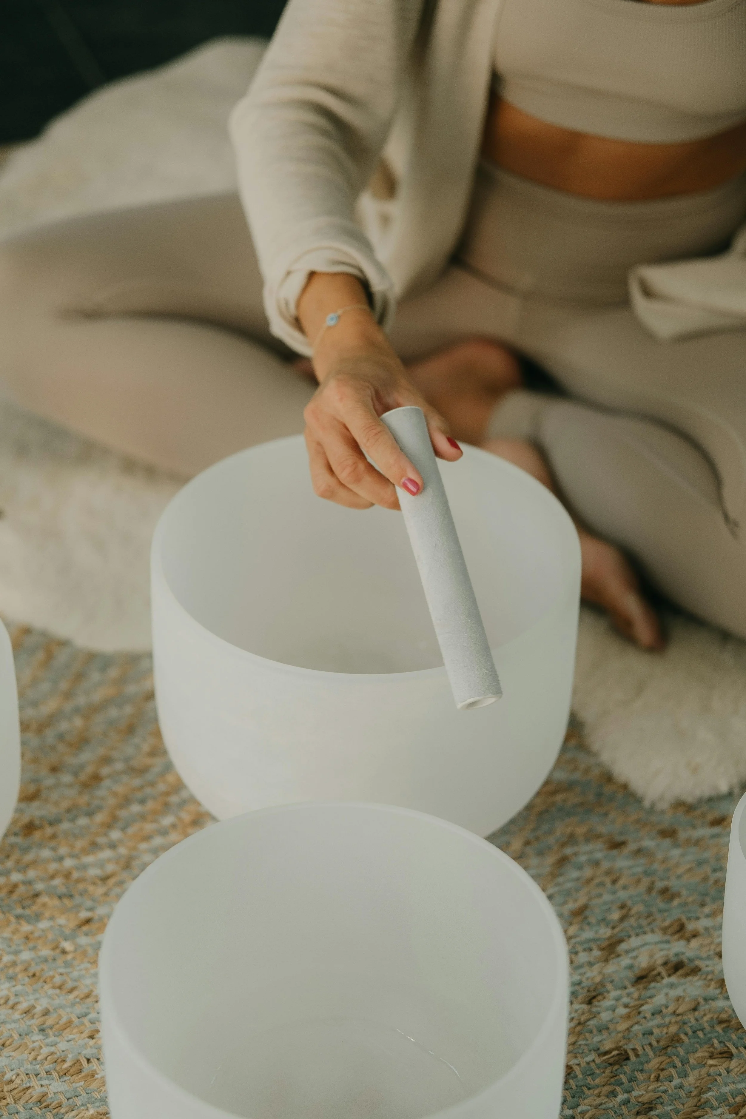 A person playing crystal singing bowls, holding a mallet, in a seated position on a rug.