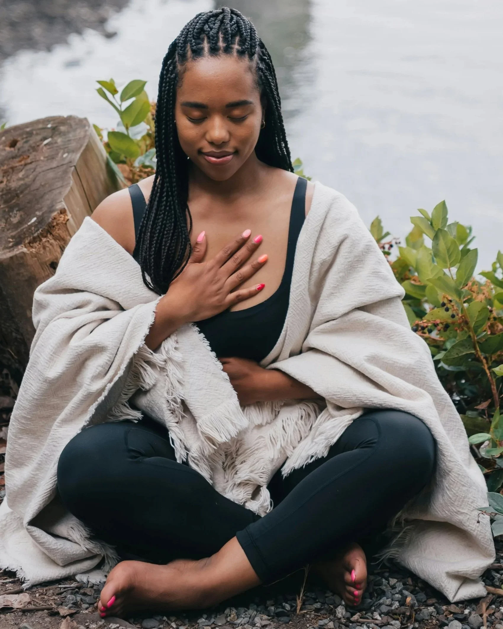 A woman sitting cross-legged outdoors near water, with her hand on her chest and eyes closed, surrounded by greenery.