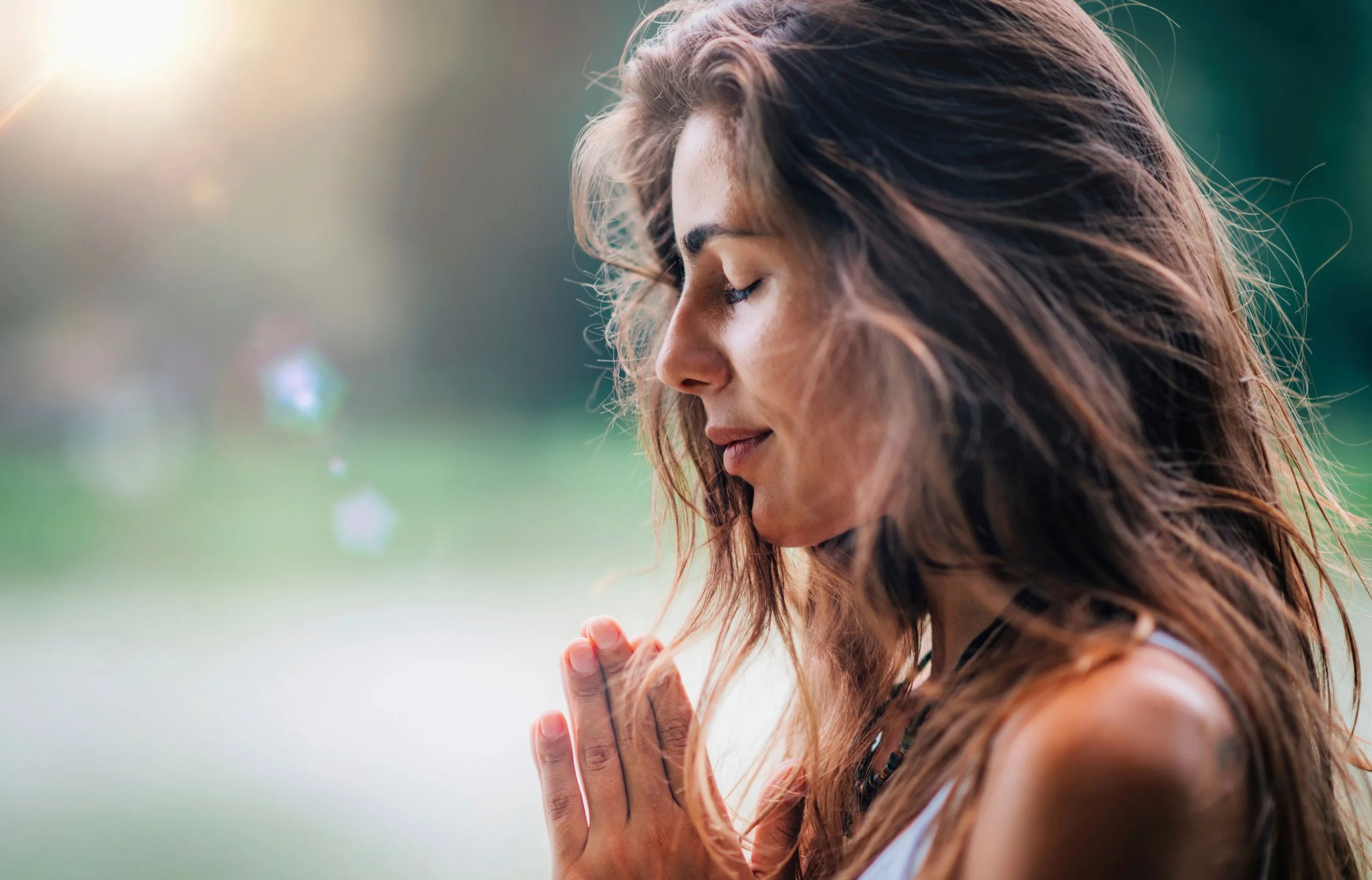 A woman with long, wavy brown hair is shown with her eyes closed, praying outdoors with her hands together, in a serene setting bathed in soft sunlight.