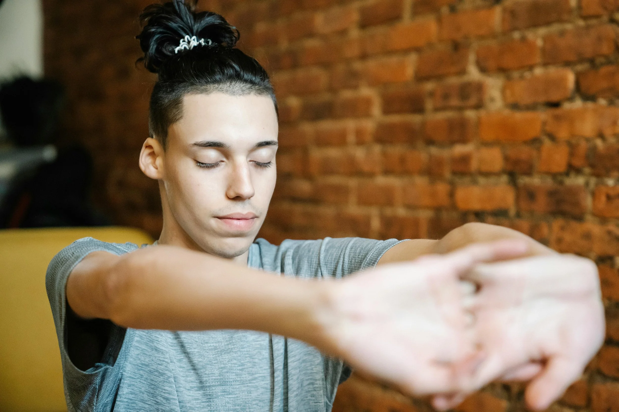 A person with short black hair in a top knot is sitting with eyes closed, wearing a gray shirt, with a brick wall background.