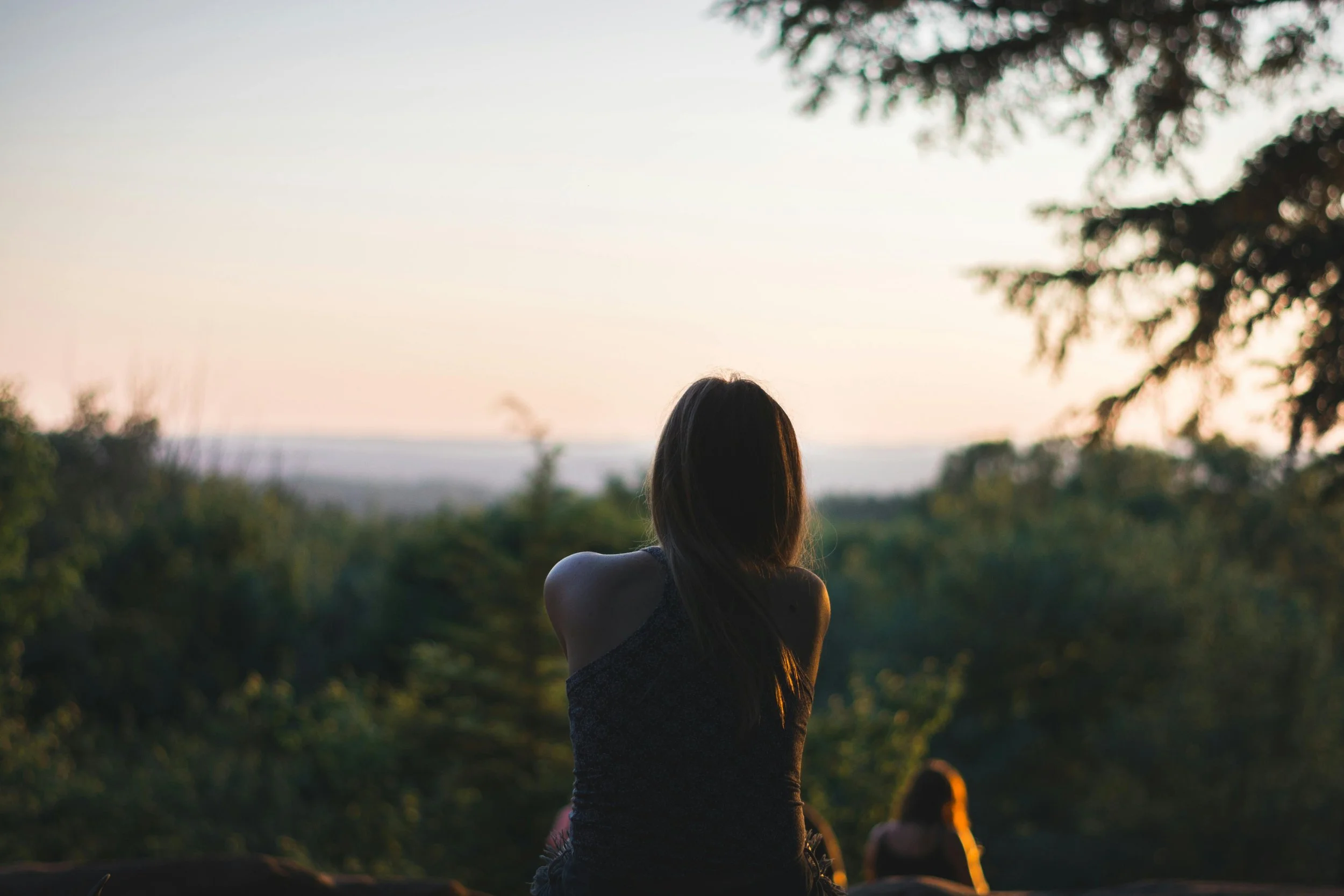A person with long dark hair sitting outdoors during sunset, facing a scenic view with trees and a blurred horizon.