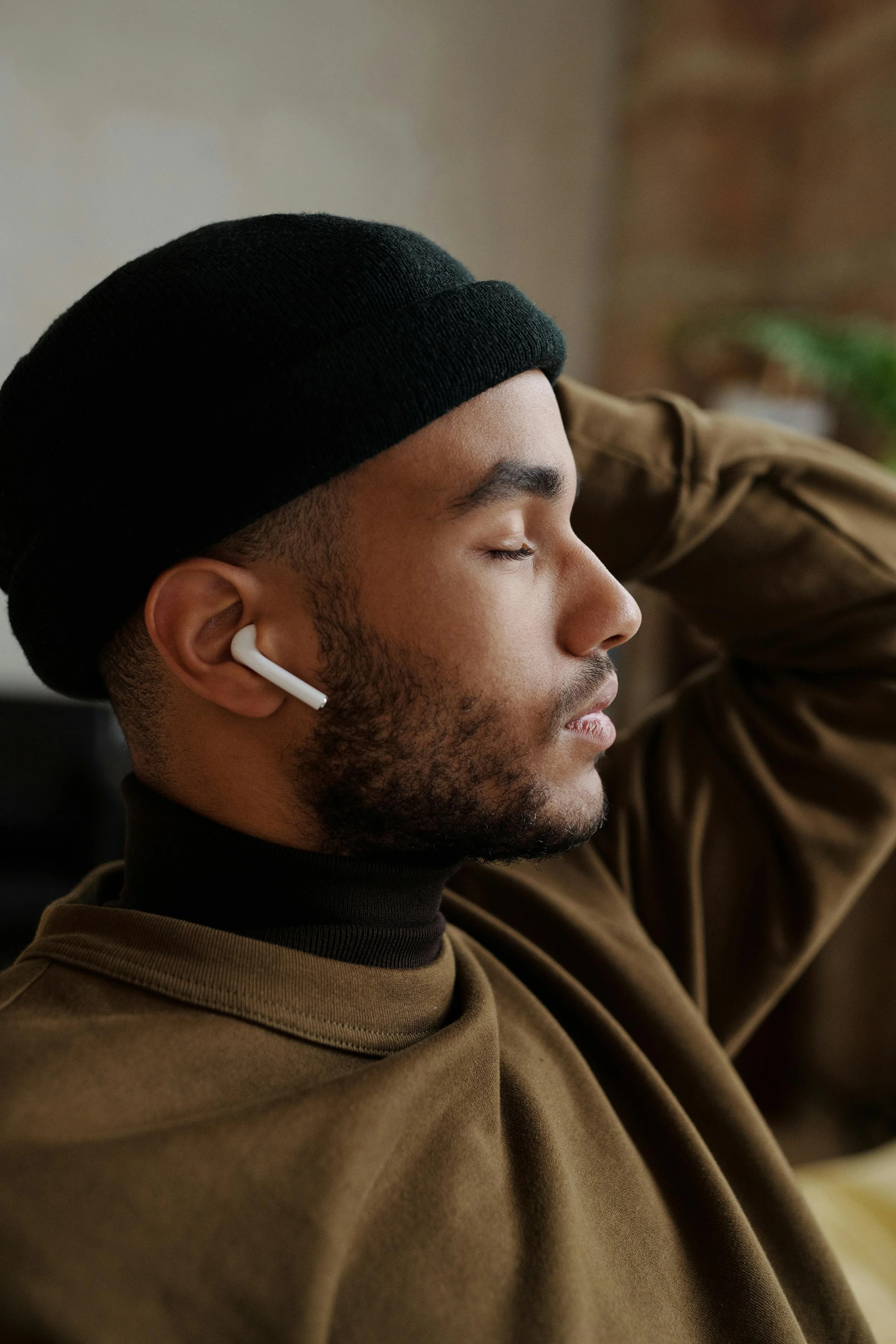 A young man with dark hair and a beard, wearing a black beanie and AirPods, relaxed with eyes closed, resting his hand on his head.