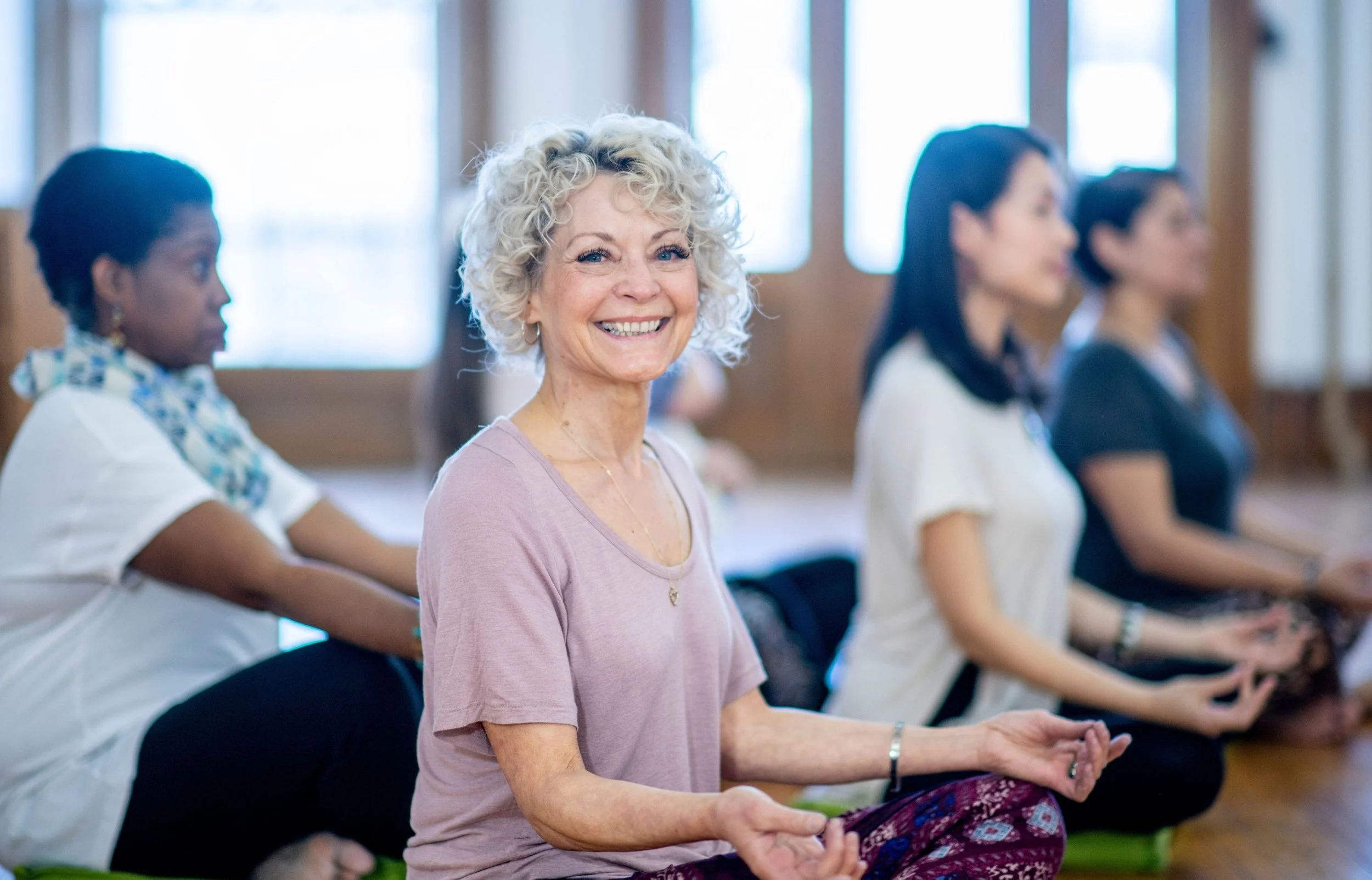 A group of women practicing yoga or meditation indoors, focusing on a smiling older woman with curly gray hair in the foreground, with others sitting in a meditation pose behind her.