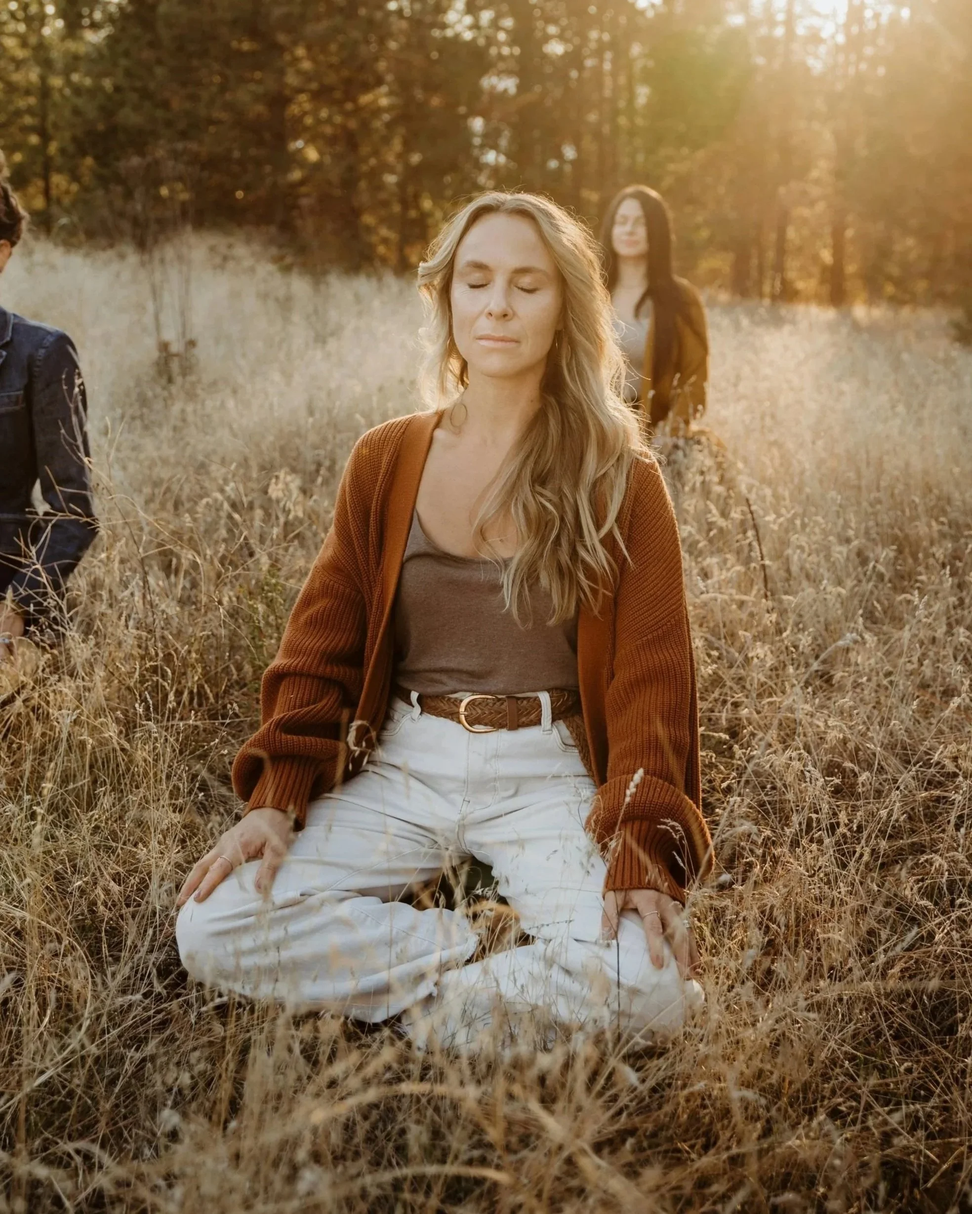 A woman with long wavy blonde hair, wearing a brown cardigan and white pants, sitting in a field of dry grass with her eyes closed. Two other women are visible in the background among trees, one with black hair and the other partially visible. The sunlight creates a warm glow over the scene.