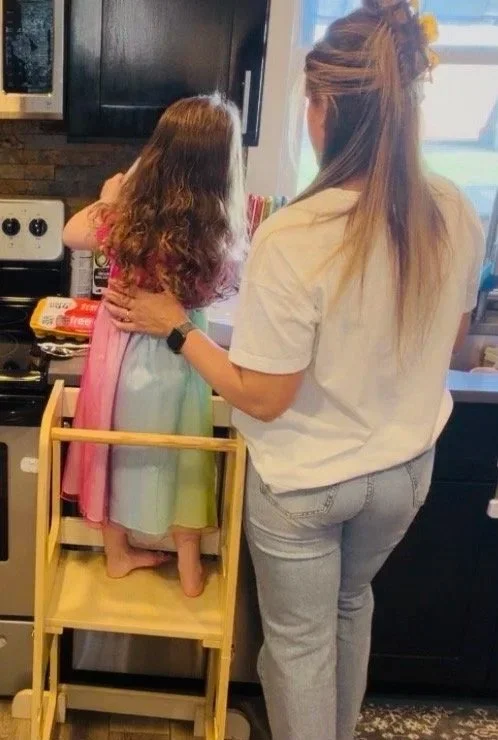 A woman helping a young girl in a rainbow-colored dress stand on a wooden step stool at a kitchen counter.