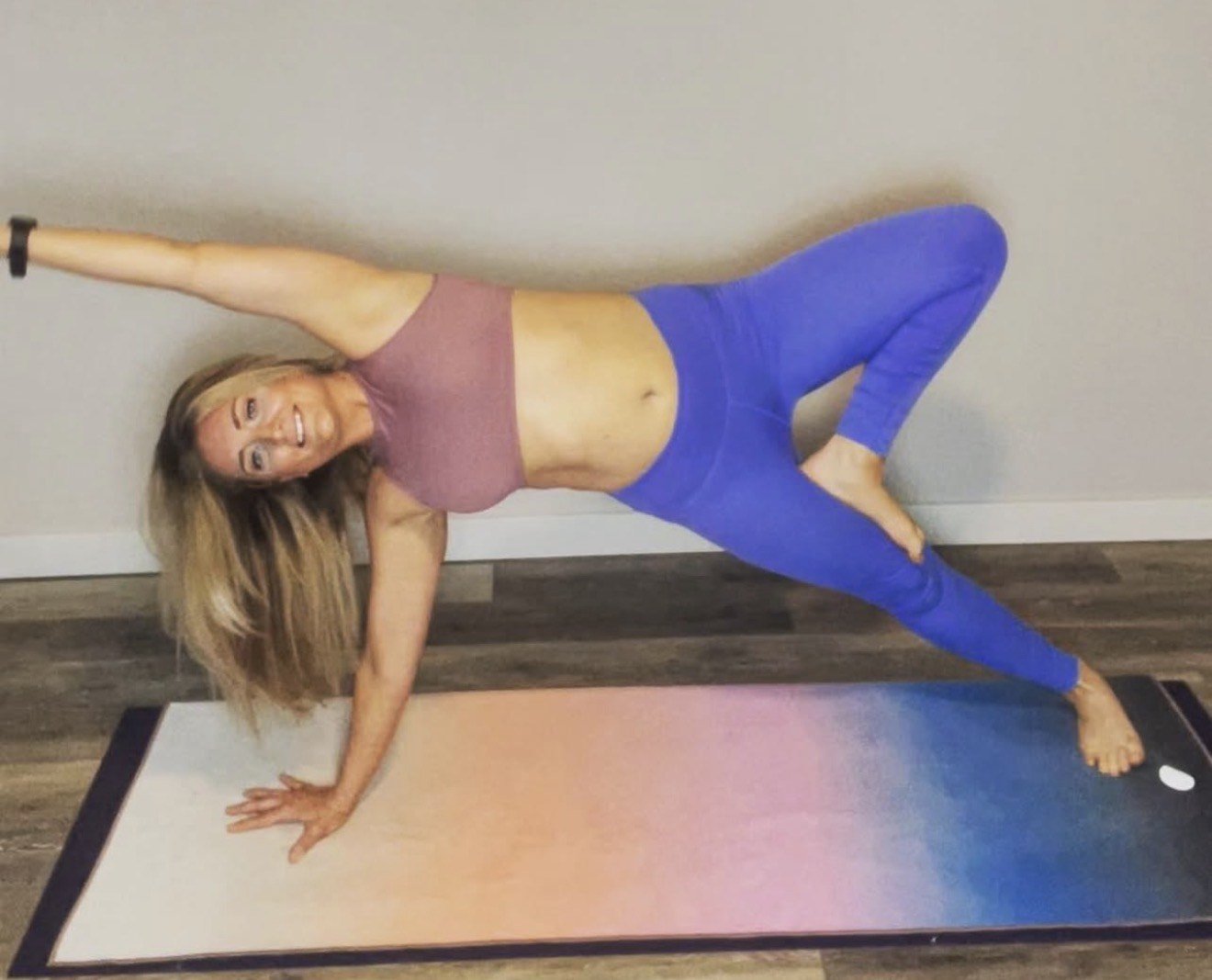 A woman with long blonde hair doing a yoga pose on a colorful yoga mat against a plain wall, wearing a mauve sports bra and blue leggings, smiling at the camera.