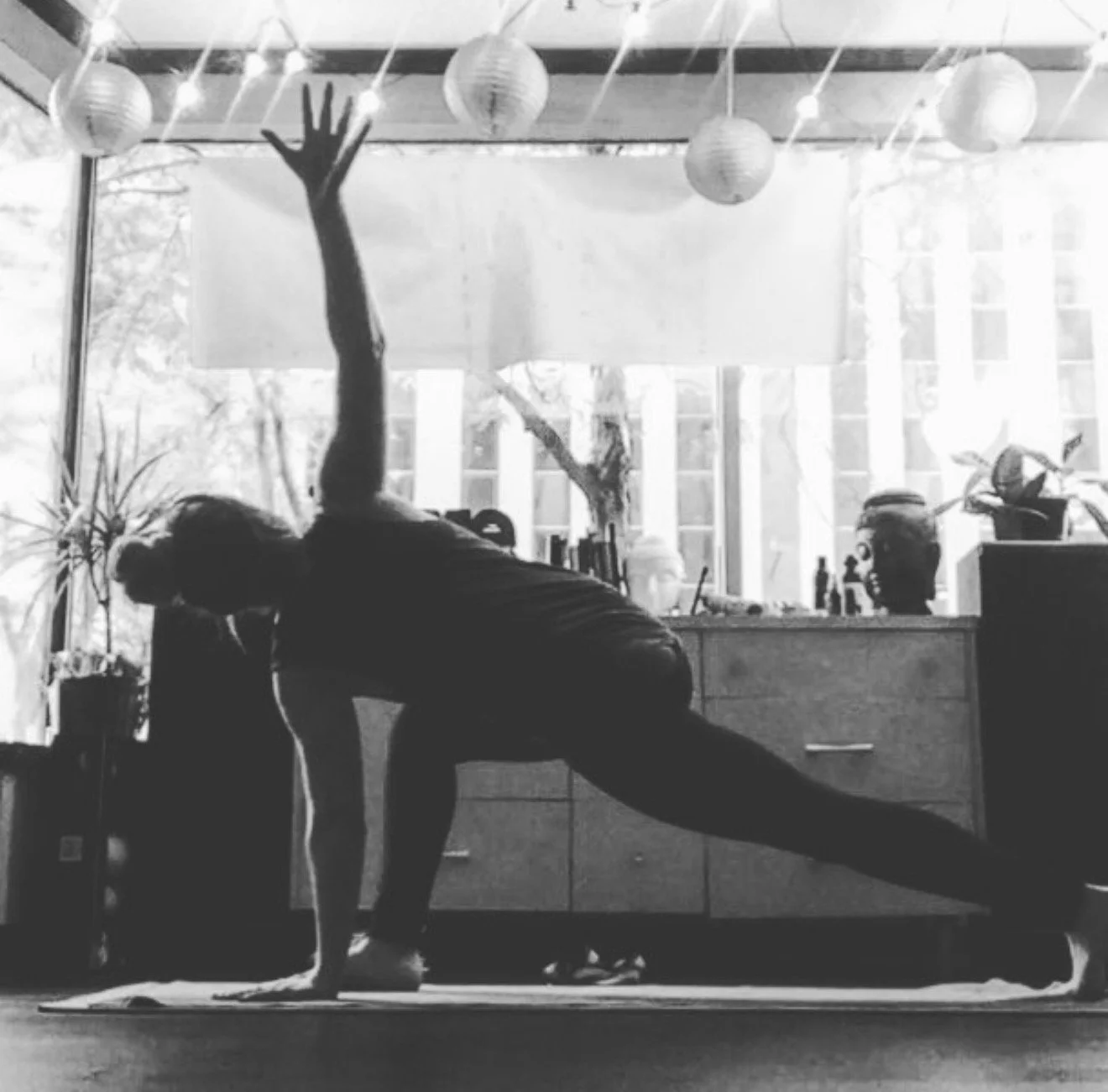 A person in a yoga pose in a room with large windows, plants, and hanging paper lanterns.