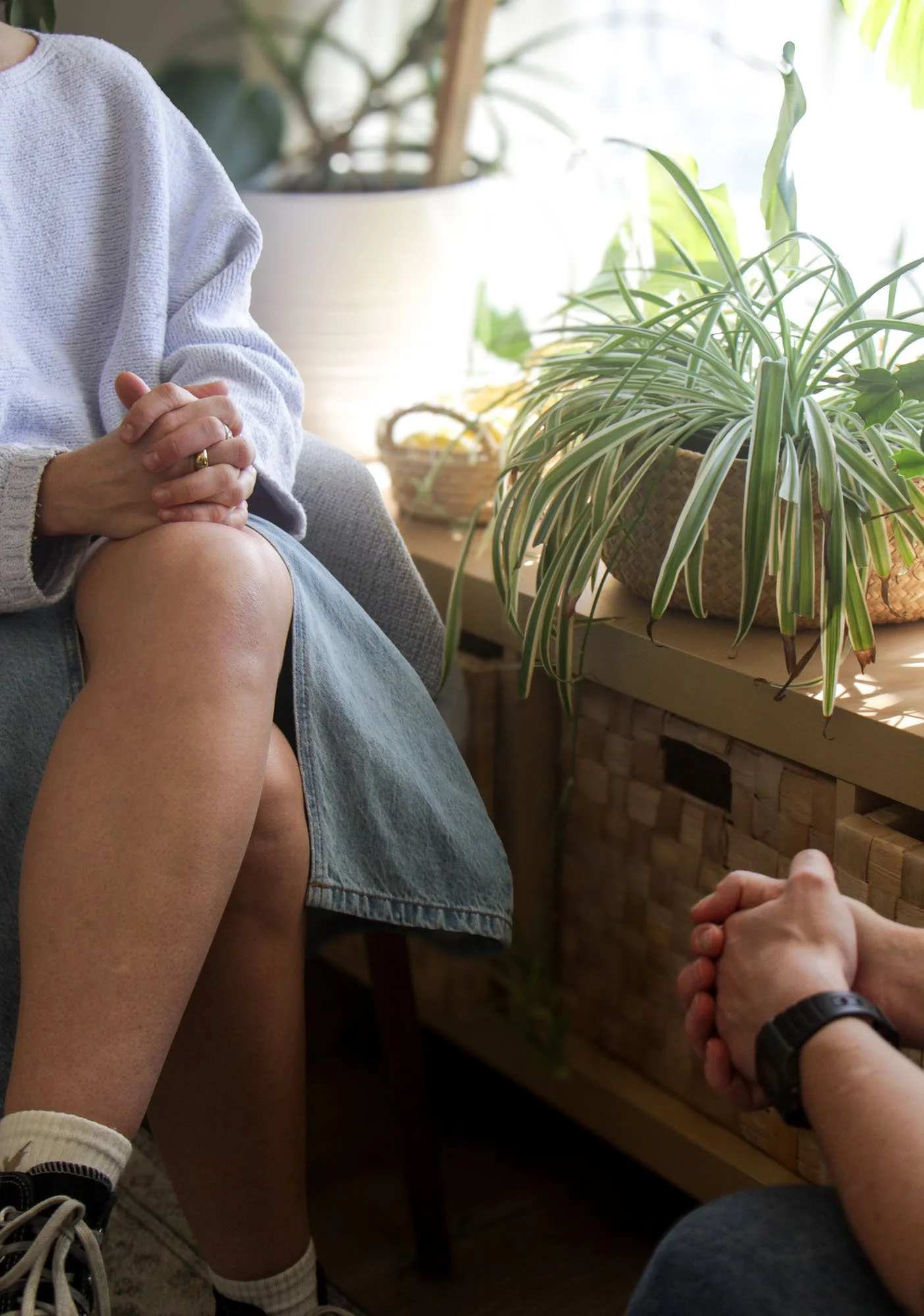 Two people sitting indoors, one with hands clasped and the other with hands folded, near a table with potted plants in a well-lit room.